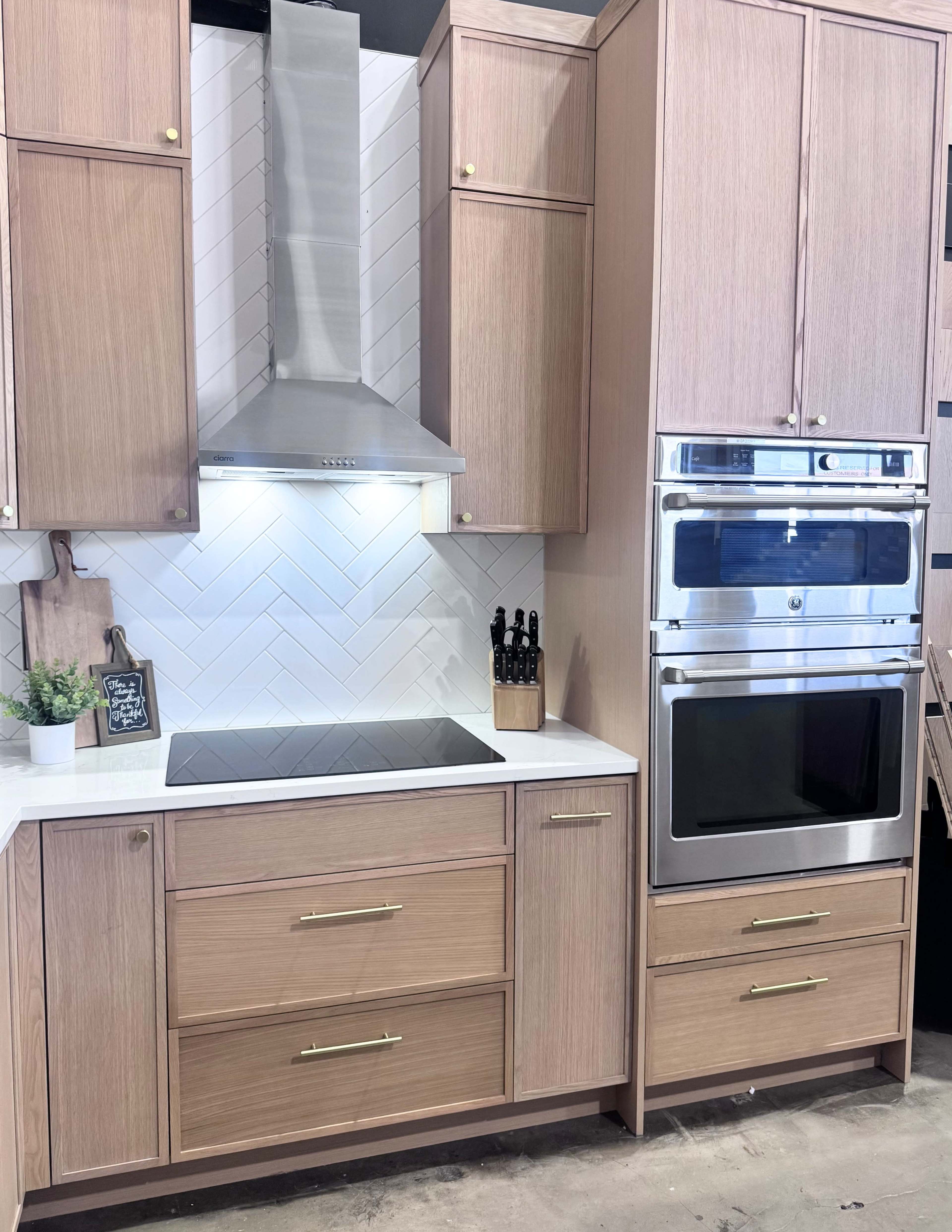 The image shows a modern kitchen featuring wooden cabinets, a stainless steel range hood, an induction cooktop, and a built-in oven stacked above a microwave.