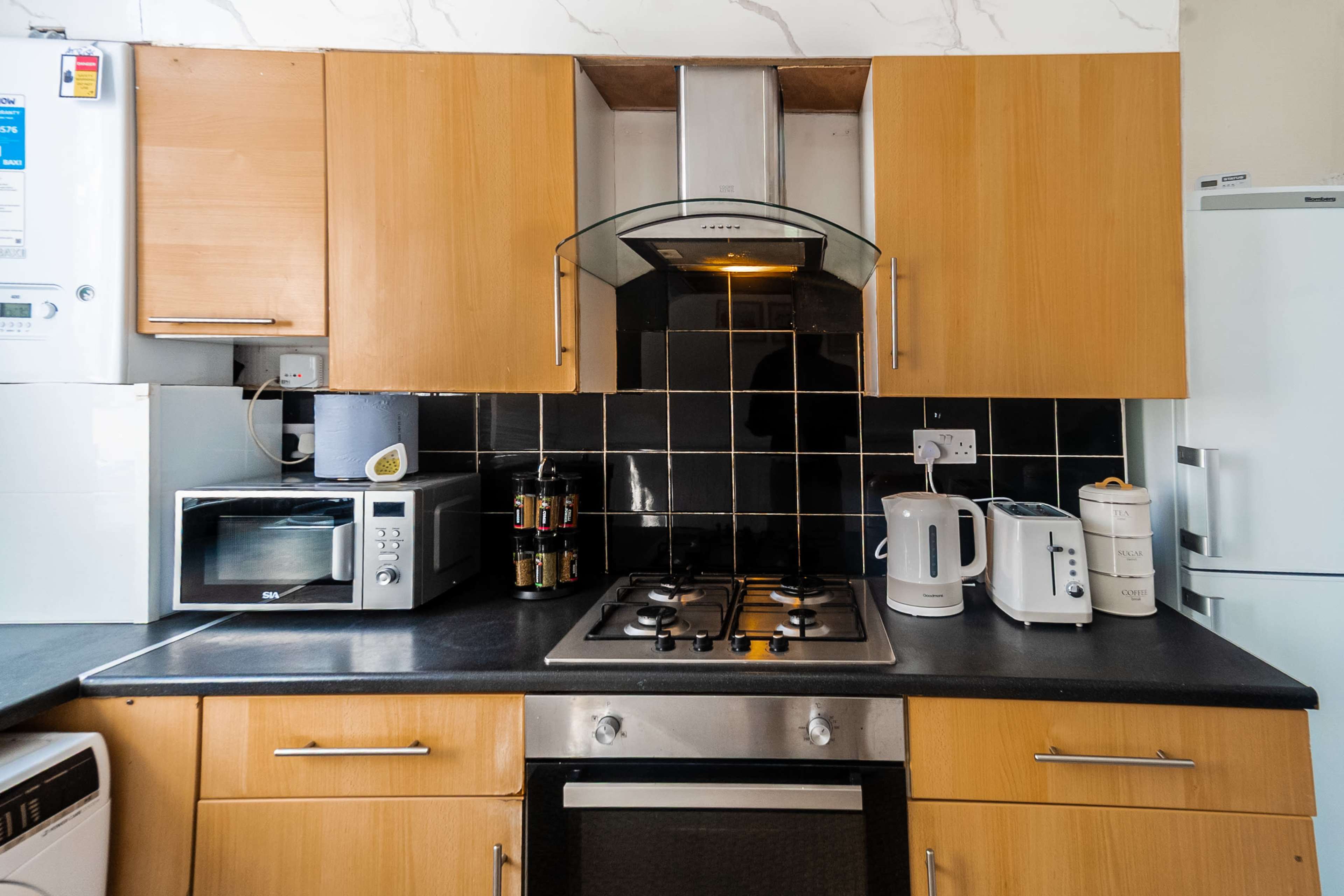 The image shows a kitchen with wooden cabinets, a stainless steel stove, a microwave, a kettle, and a refrigerator, all against a black tiled backsplash.