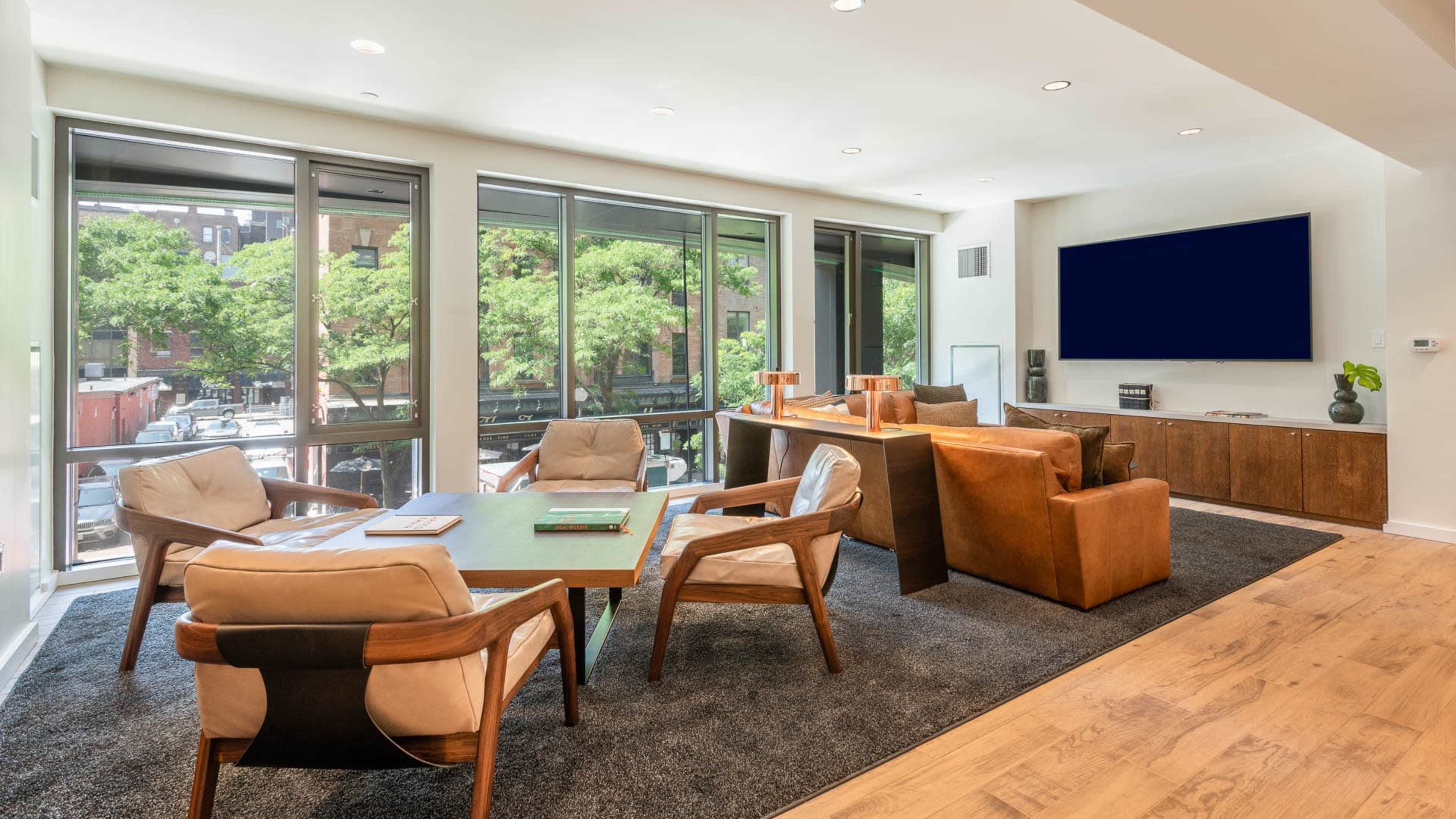 The image shows a modern living room with large windows, a wooden coffee table, four chairs, and a media unit along one wall.