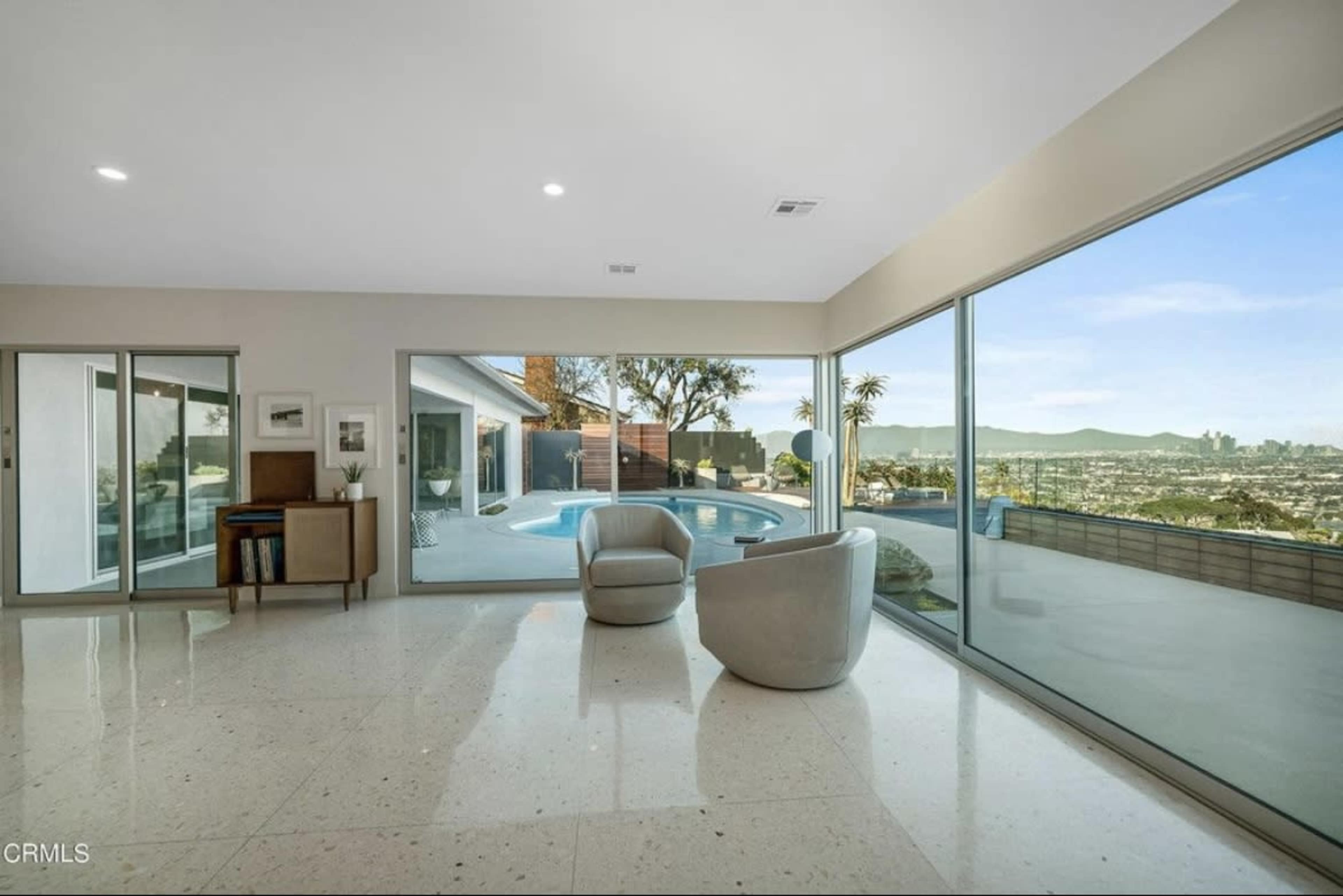 A modern living room features large glass windows overlooking a pool and a panoramic city view.