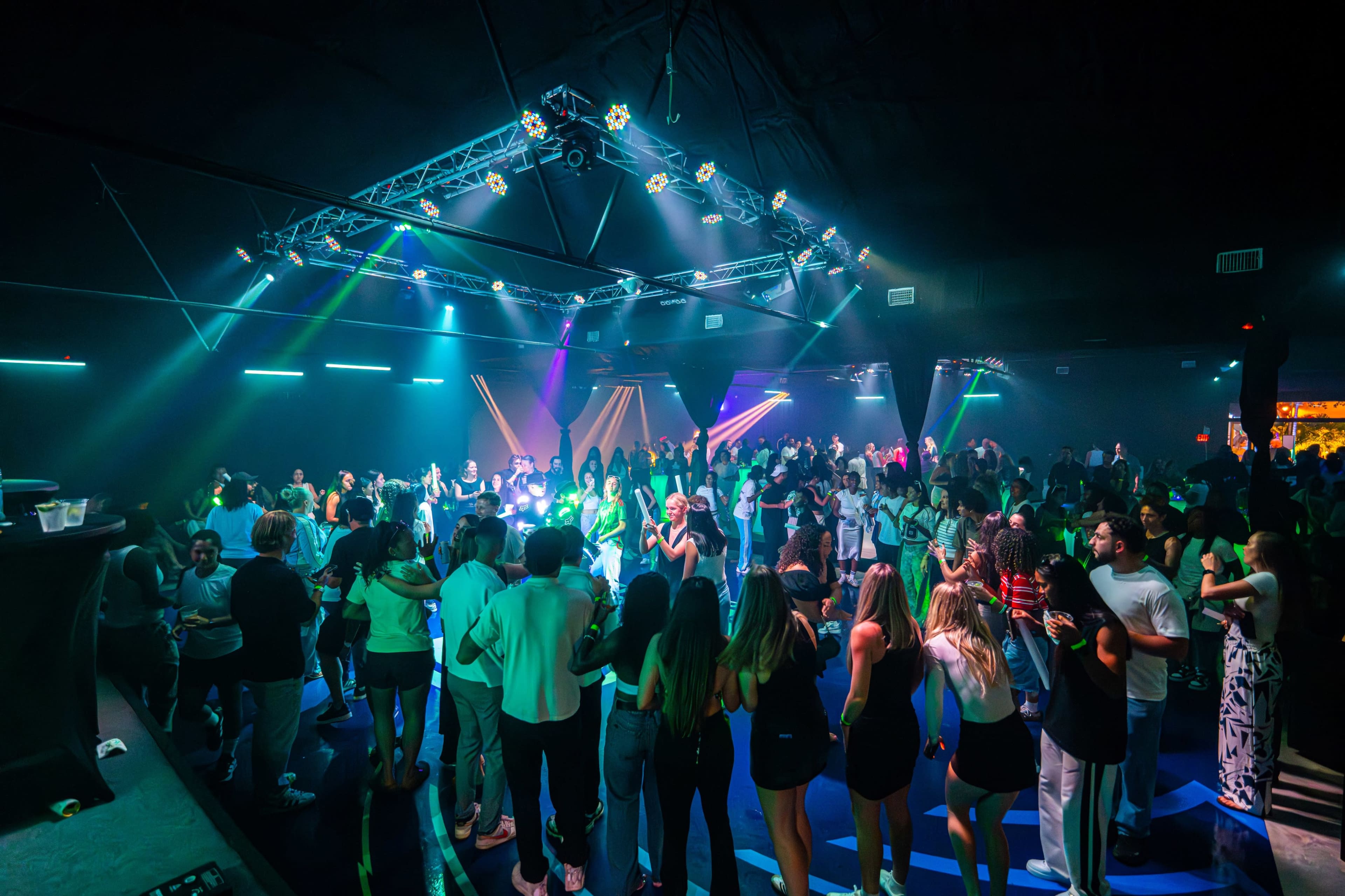 A large crowd dances under colorful lights in a lively nightclub.