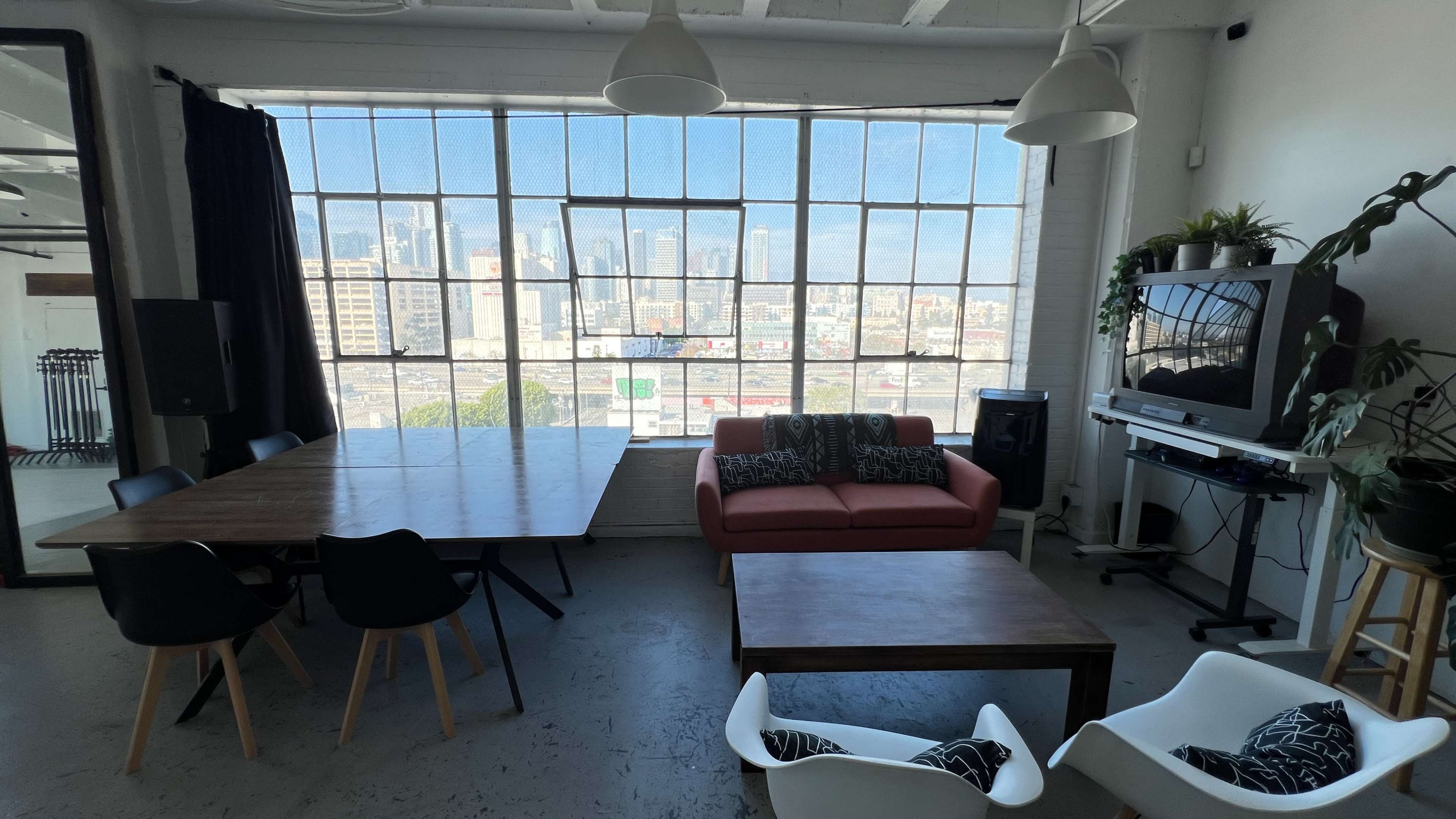 The image shows a modern, bright room with large windows overlooking a cityscape, featuring a wooden dining table, a red couch, and several chairs.