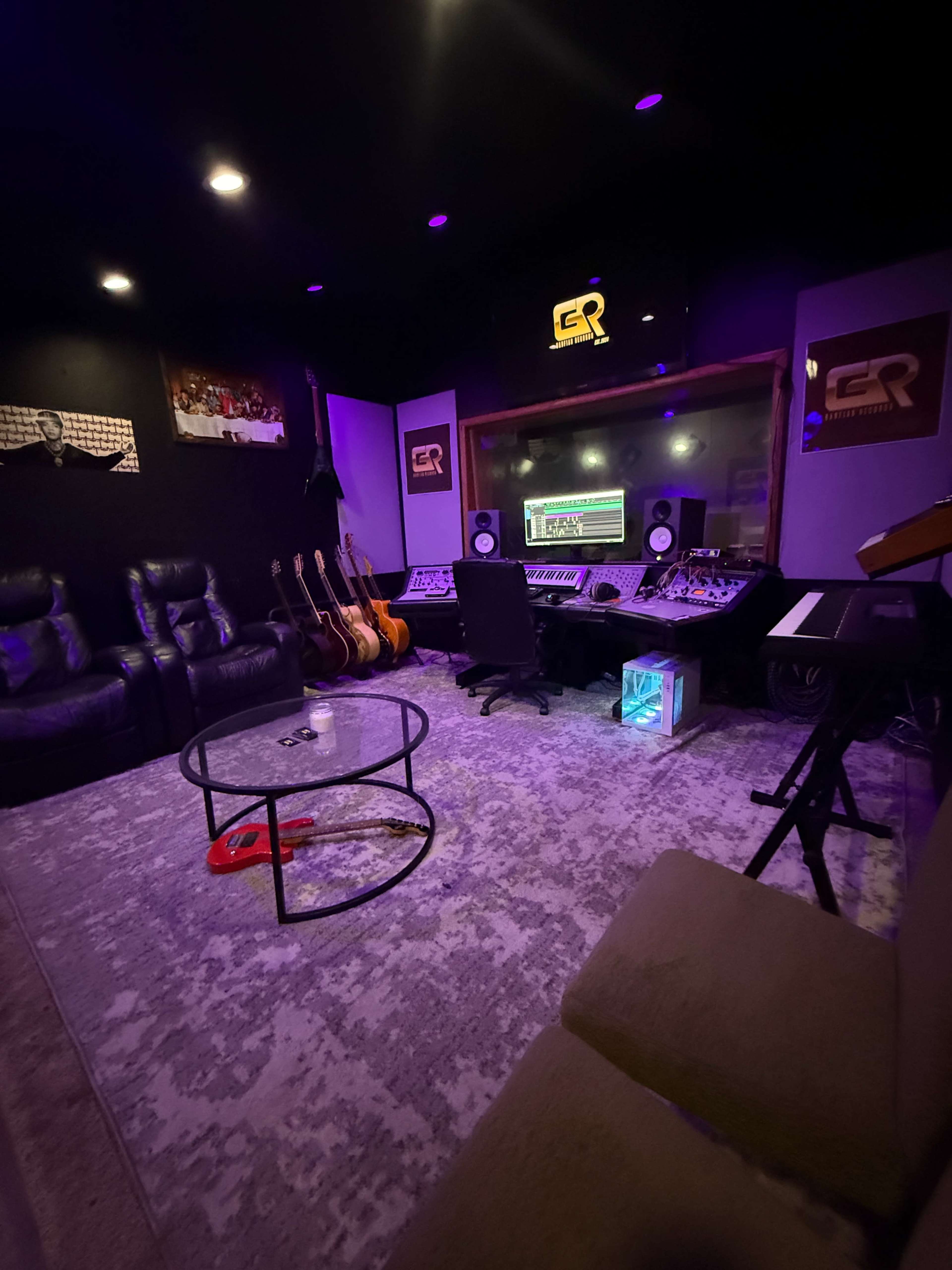 The image shows a music recording studio featuring a control desk with sound equipment, guitars on stands, and a cozy seating area with a circular glass coffee table.