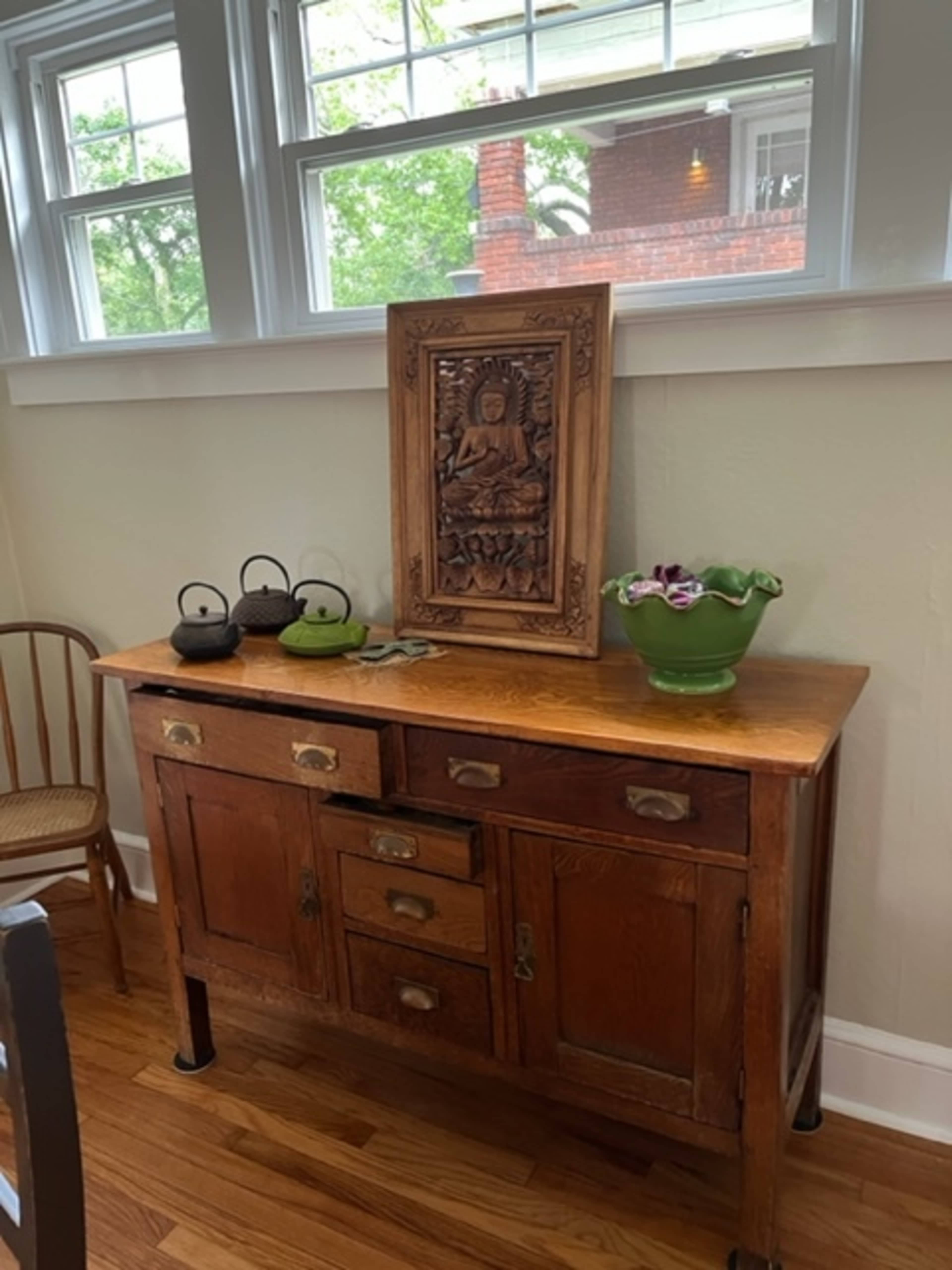 A wooden sideboard displays a carved wooden panel and a green glass bowl, with two teapots positioned on top.