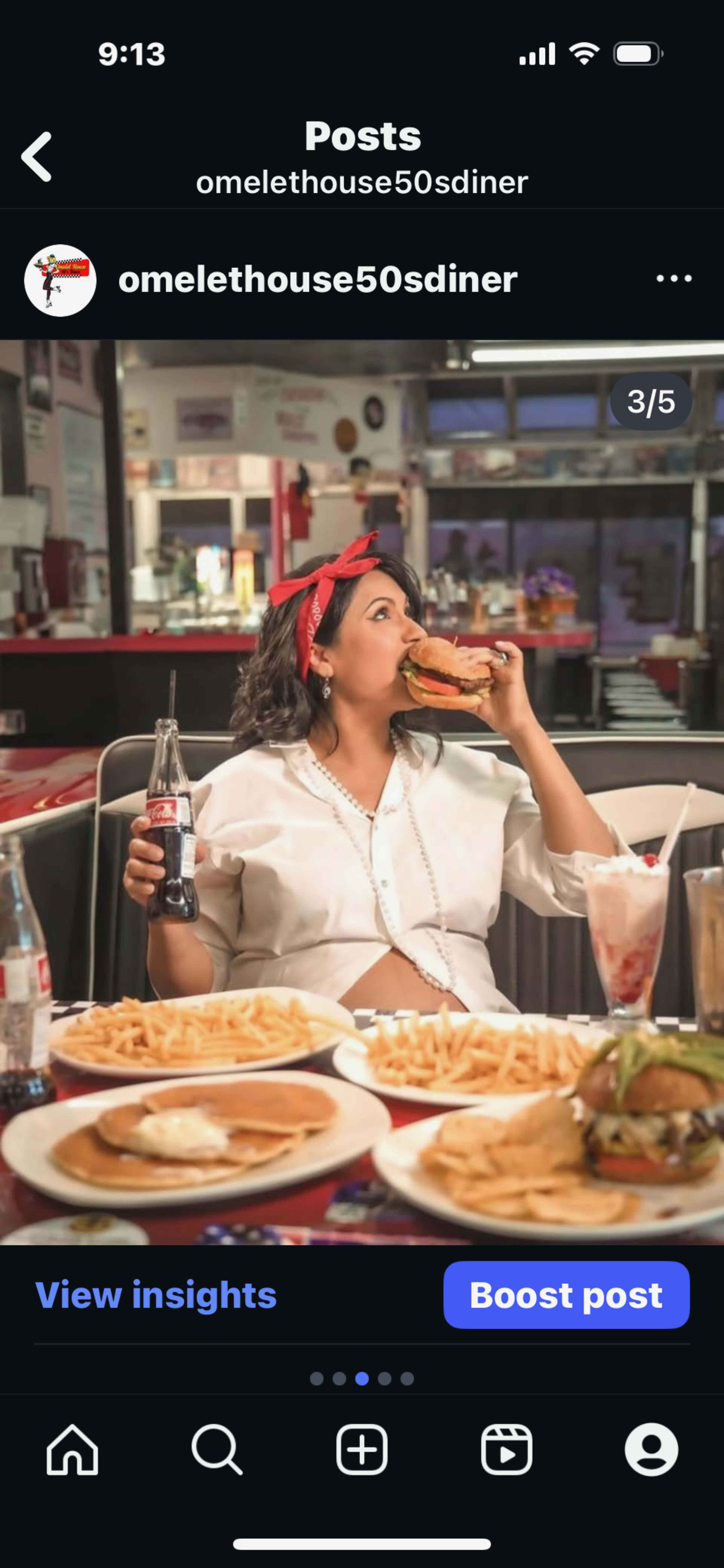 A woman wearing a red bandana takes a bite of a sandwich while seated at a diner table filled with food, including fries and a burger.