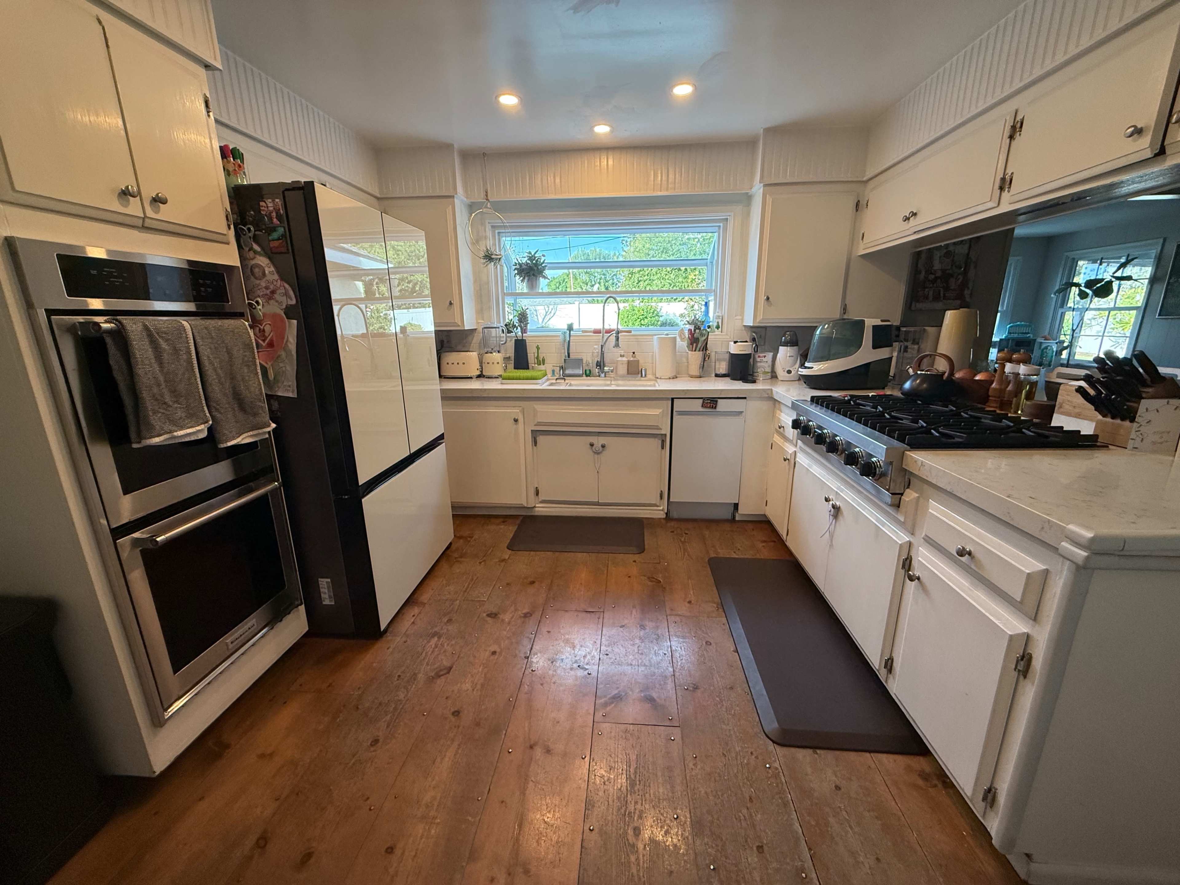 The image shows a modern kitchen equipped with stainless steel appliances, a large window above the sink, and wooden flooring.