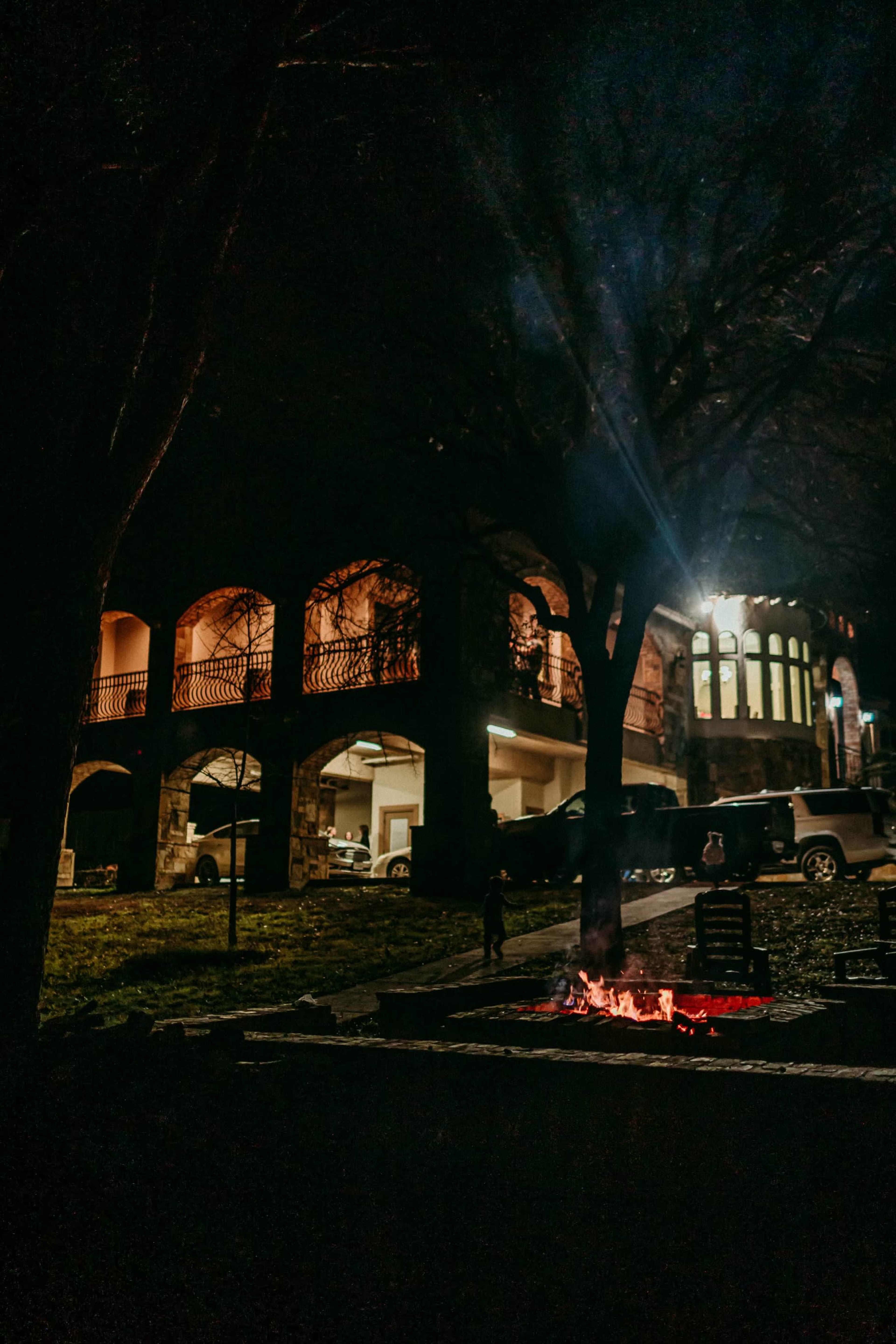 A well-lit stone building with arched balconies stands in the background, while a small fire burns in the foreground under a tree at night.