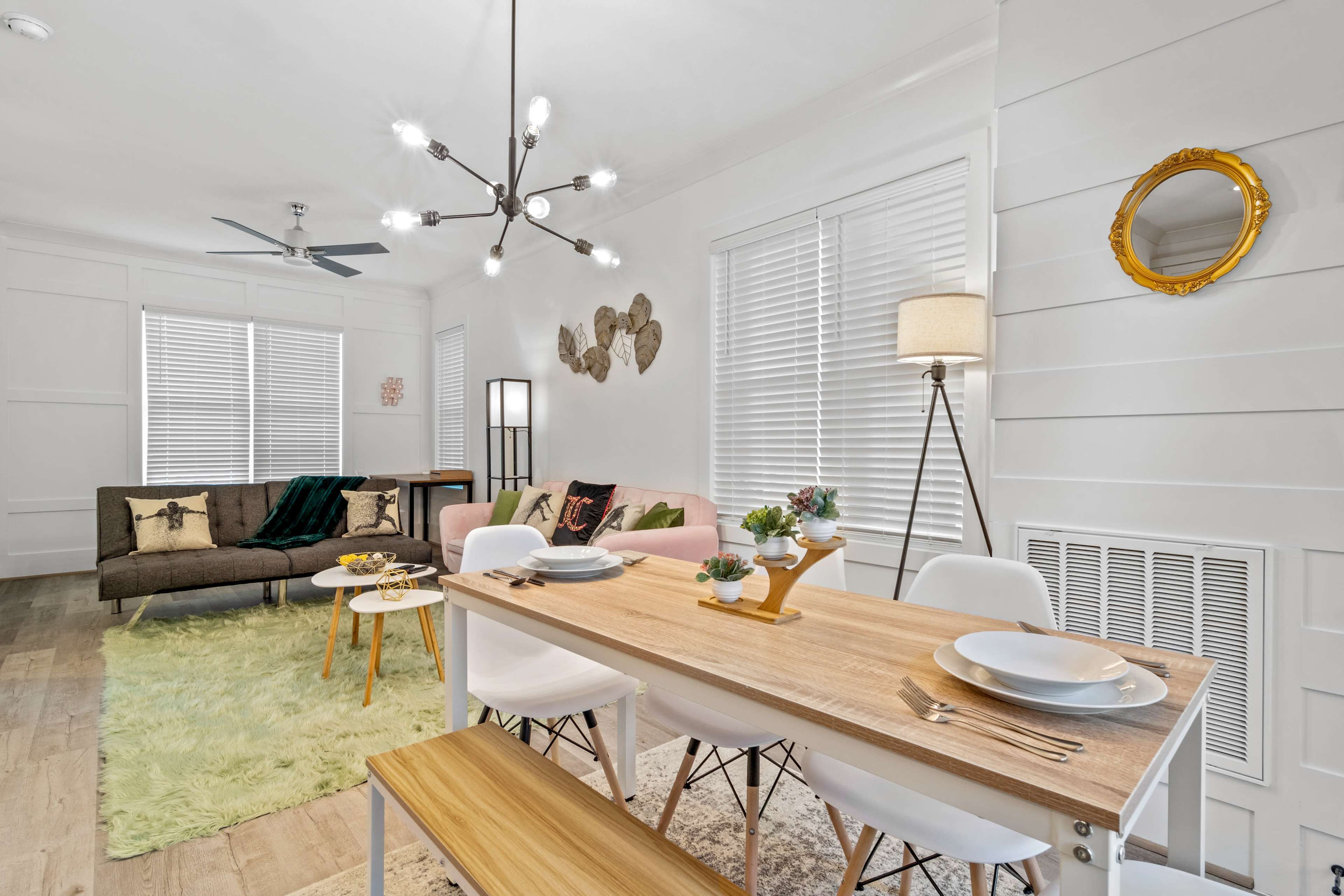 A modern living and dining area features a wooden table set for a meal, surrounded by white chairs, with a sofa, decorative items, and large windows in the background.