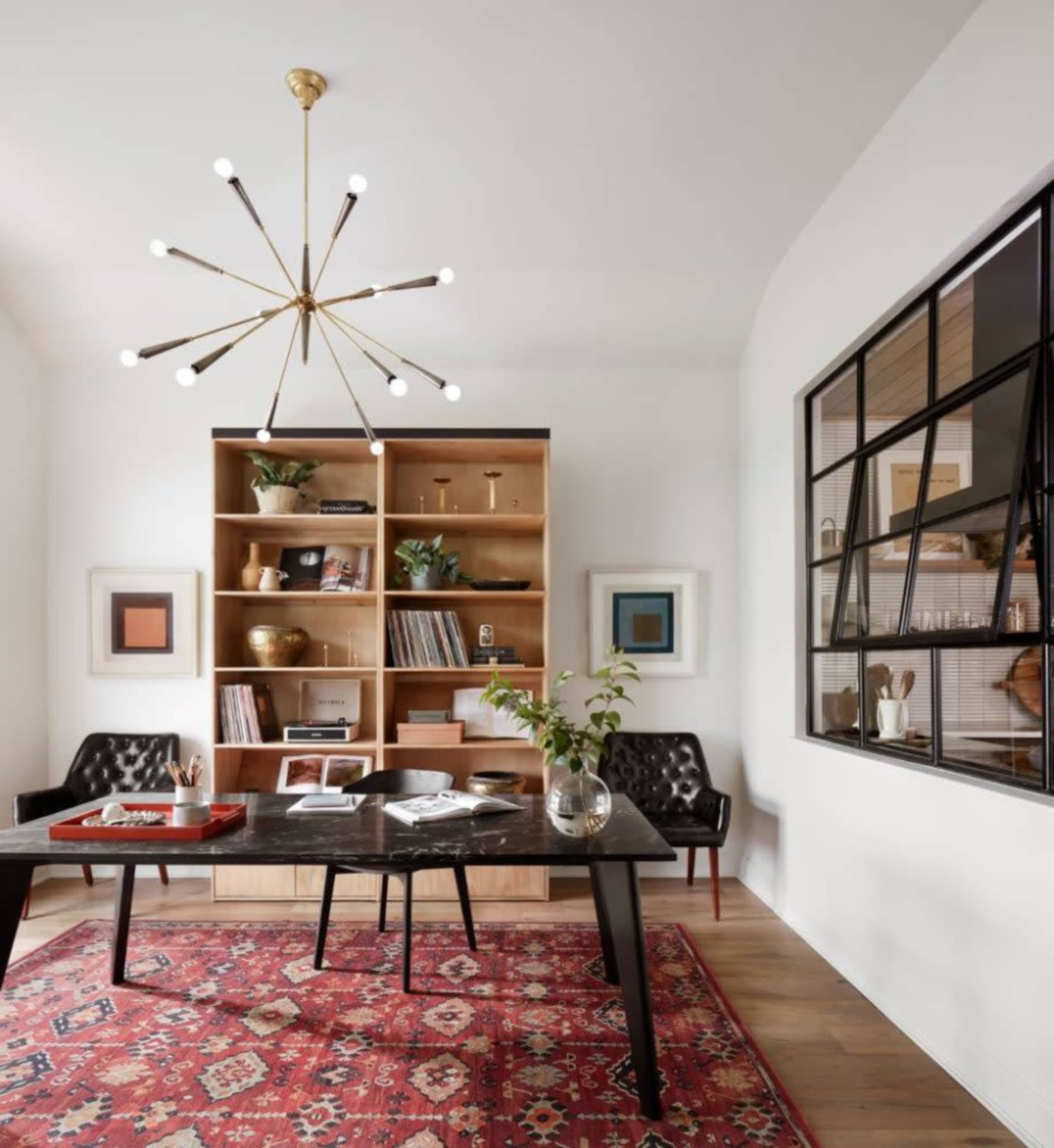 A modern office features a large dark table, a bookshelf filled with books and decorative items, and a patterned rug on the hardwood floor.
