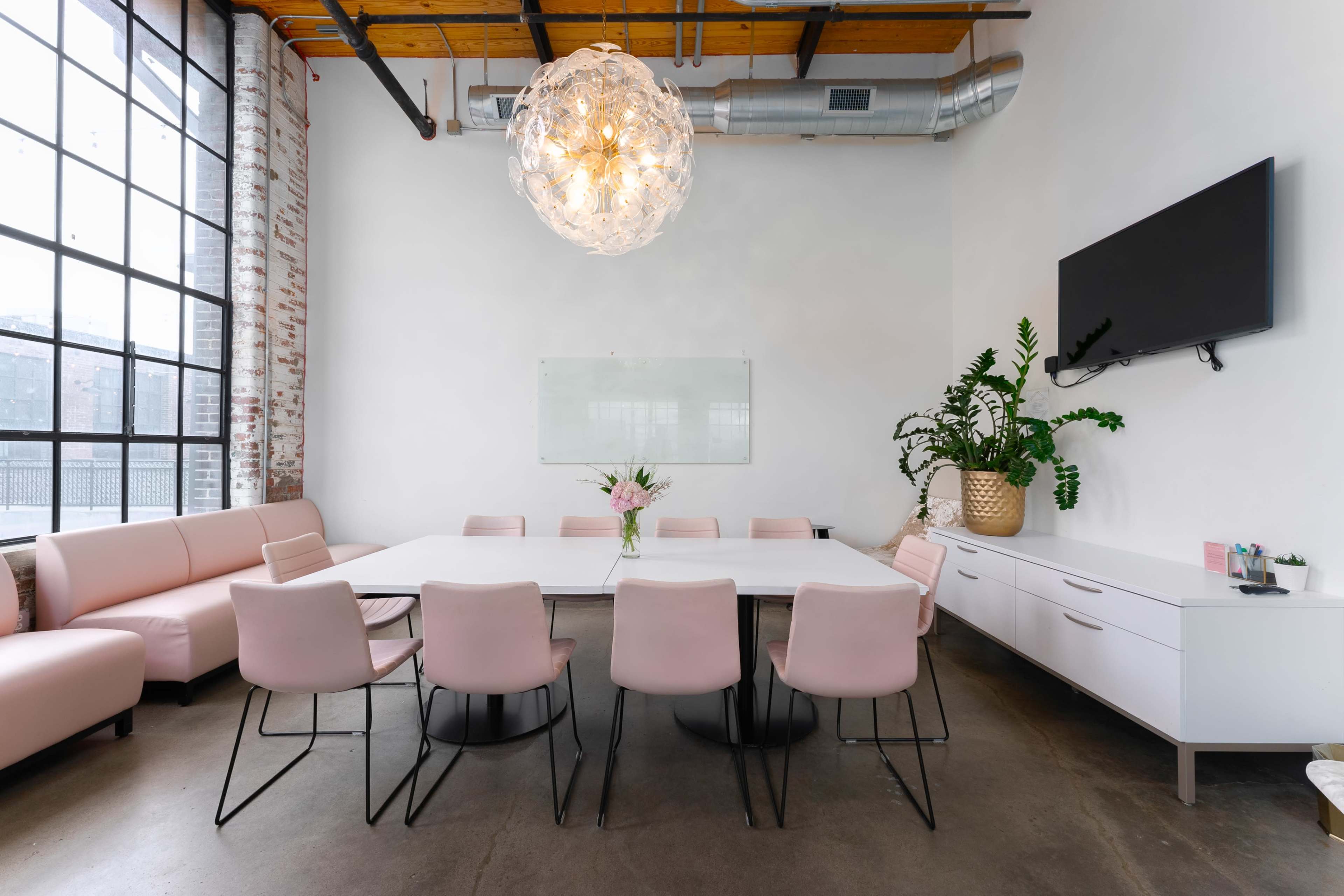 A modern meeting room features a large white table surrounded by pink chairs, illuminated by a decorative light fixture, with a plant and a whiteboard on the wall.