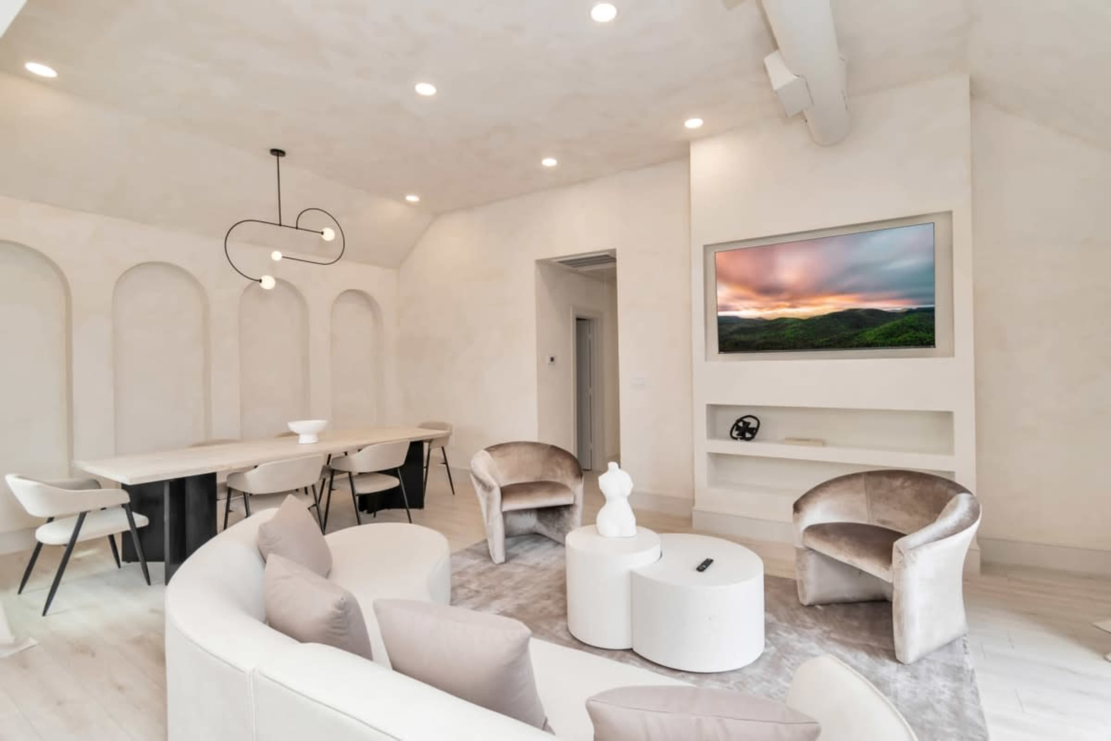 A modern living space featuring a curved sofa, two chairs, a dining table, and a wall-mounted television with a scenic backdrop.