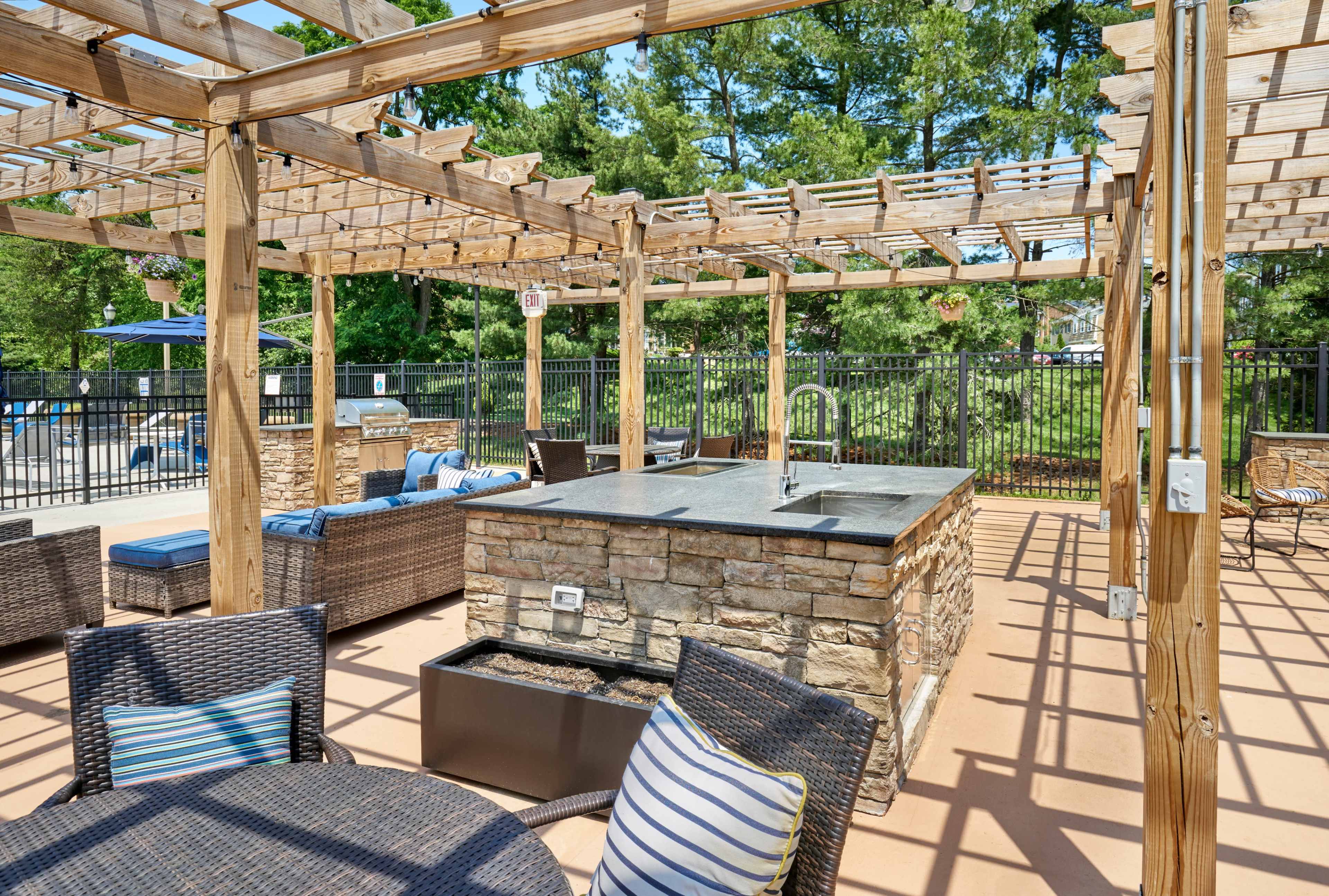 The image shows an outdoor patio area with a stone barbecue grill, seating arrangements made of wicker furniture, and a pergola structure overhead.