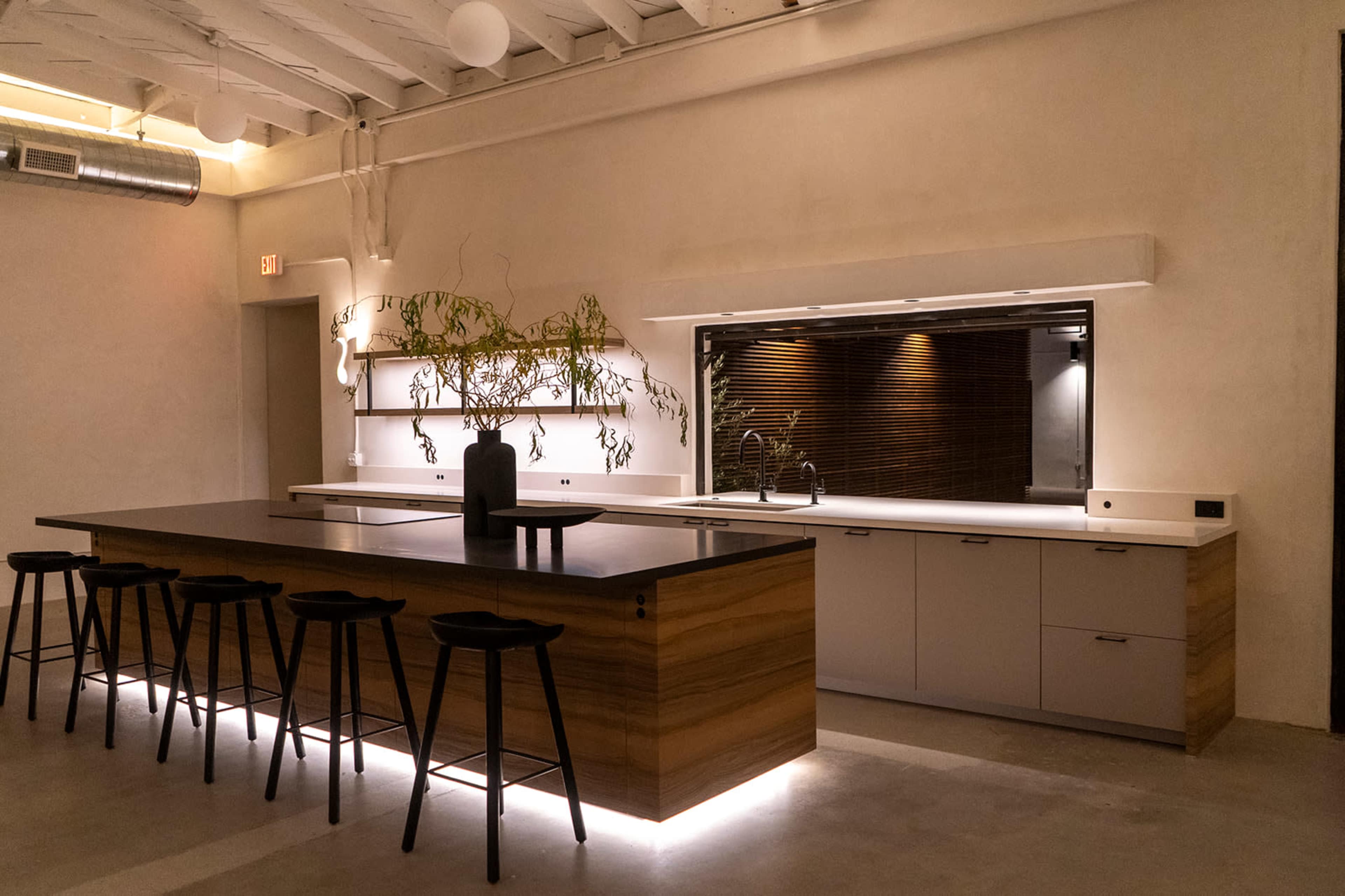 A minimalist kitchen features an island with wooden cabinets, black bar stools, and under-cabinet lighting, complemented by a simple backsplash and wall decor.