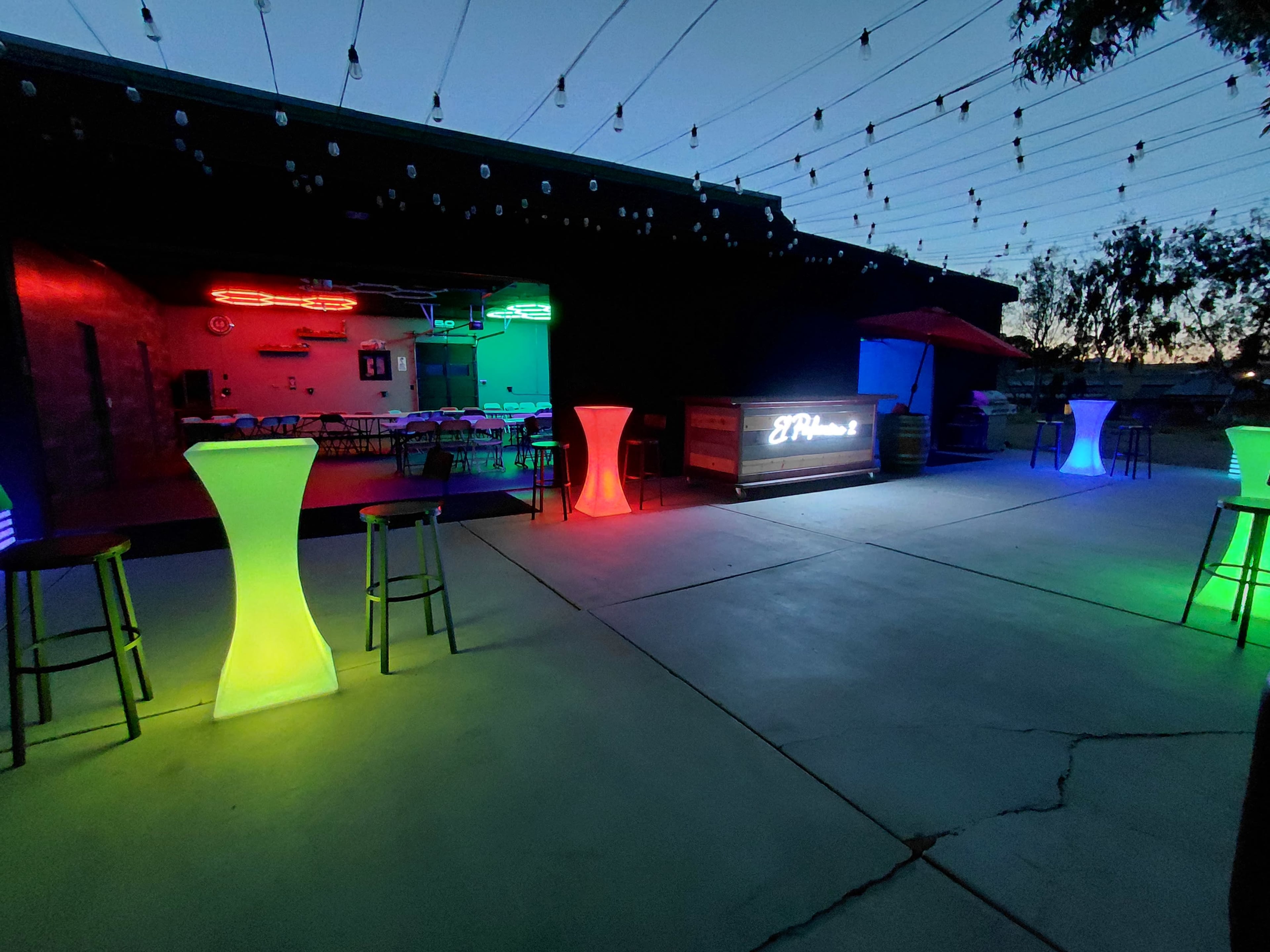 The image shows a spacious outdoor area with brightly lit tables and a bar, where colorful neon lights illuminate the scene under a twilight sky.