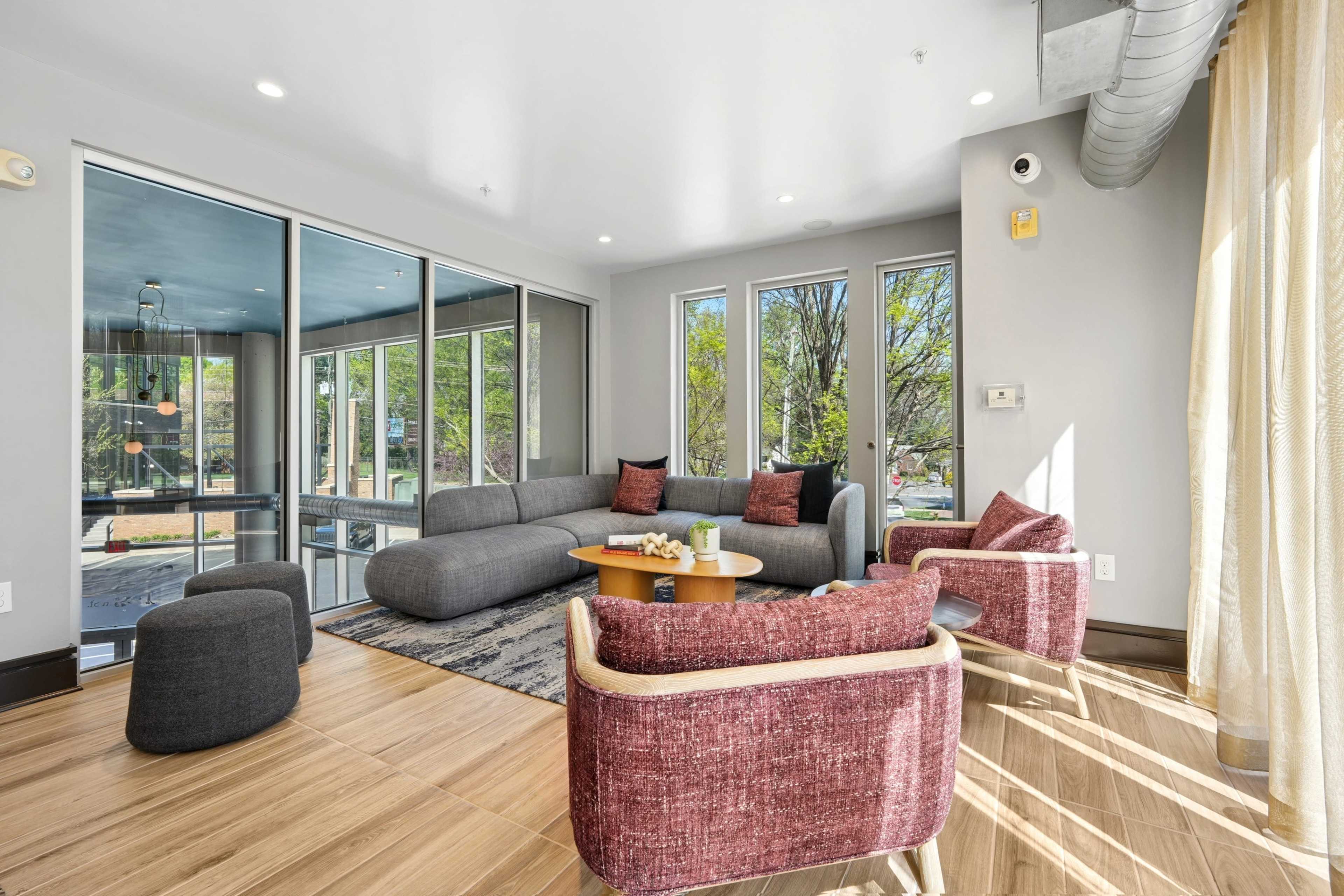 The image shows a modern living area with a gray sectional sofa, two red accent chairs, a small round table, and large windows allowing natural light.