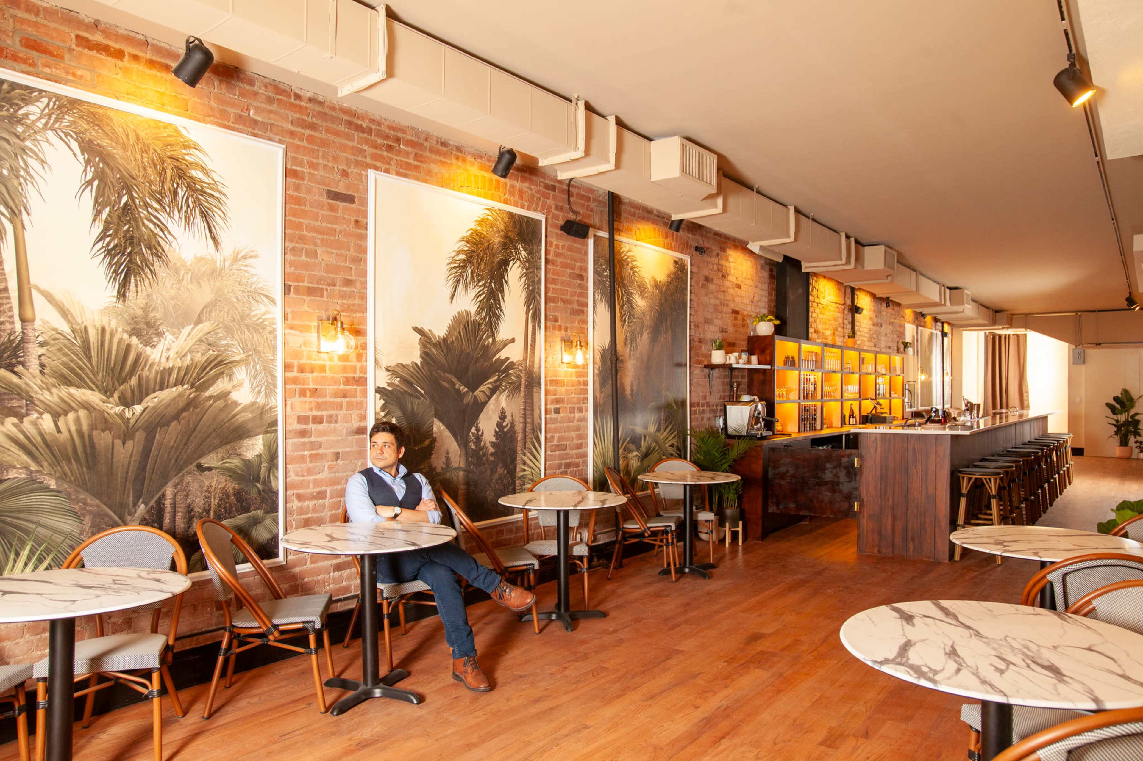 A café interior features brick walls adorned with large tropical murals, small round tables, and a bar in the background.