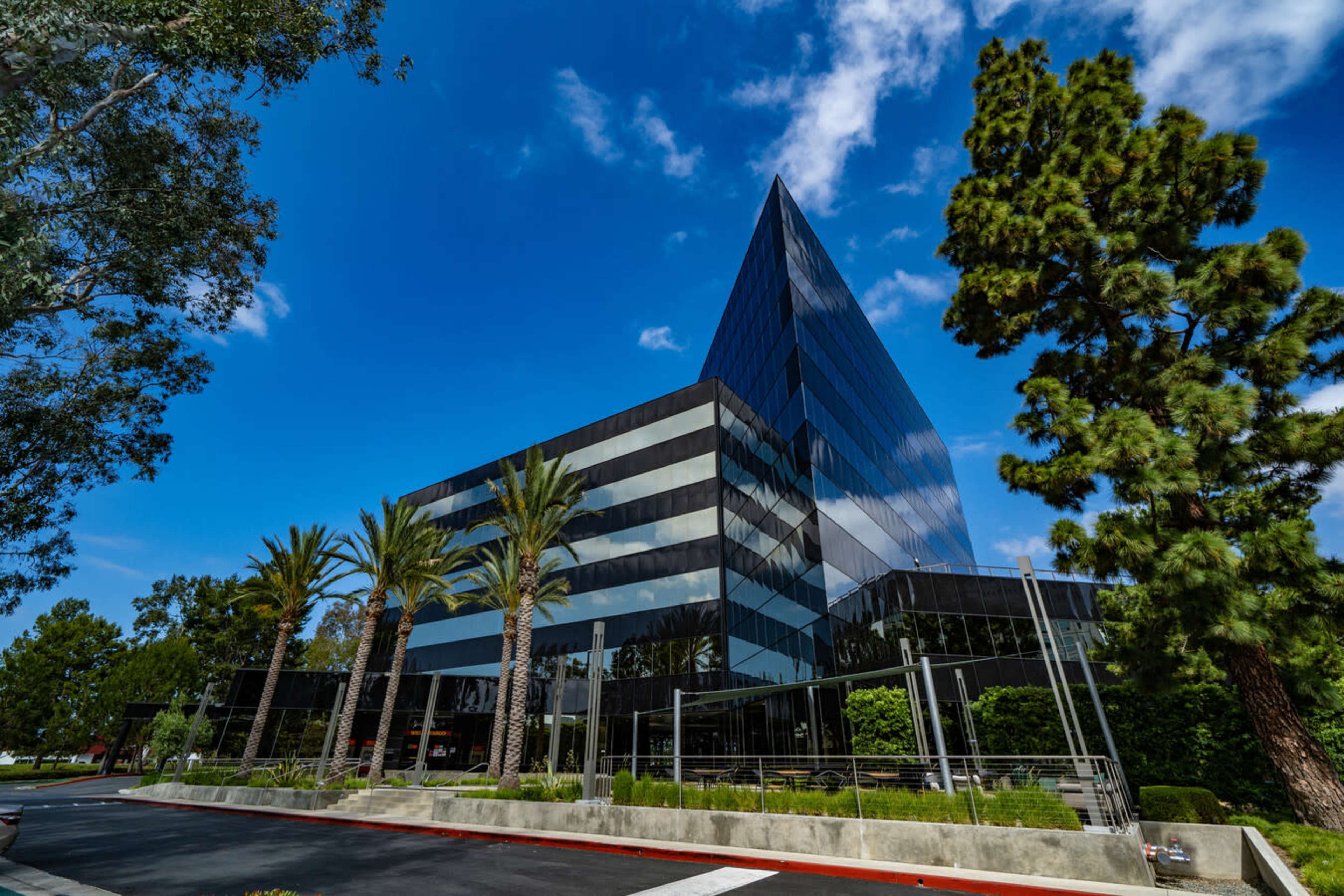 A modern, angular glass building is surrounded by palm trees and lush greenery under a partly cloudy sky.