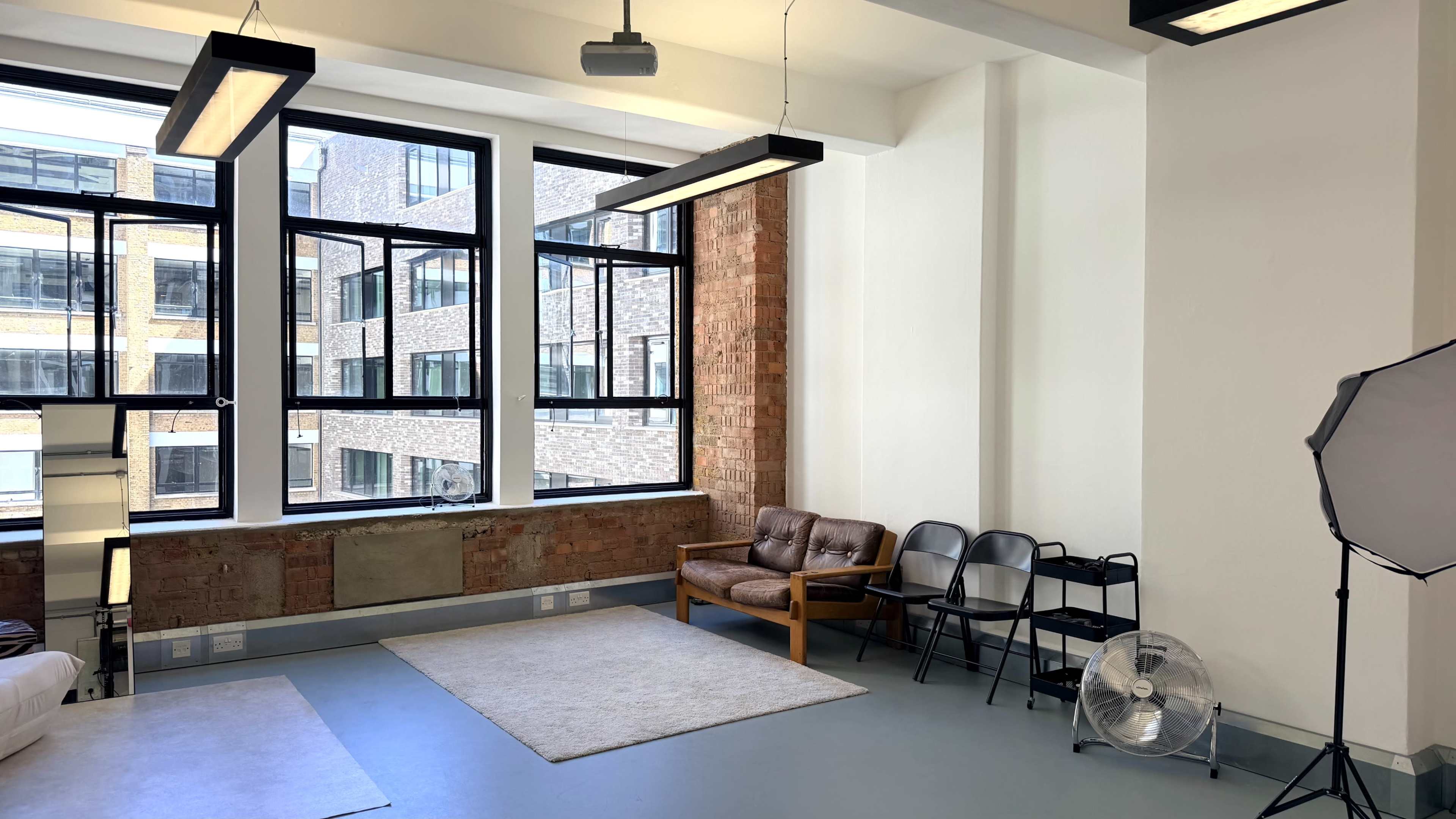 The image shows a bright, minimalist indoor space featuring large windows, a brown leather couch, two black chairs, a fan, and a light-colored rug on the floor.