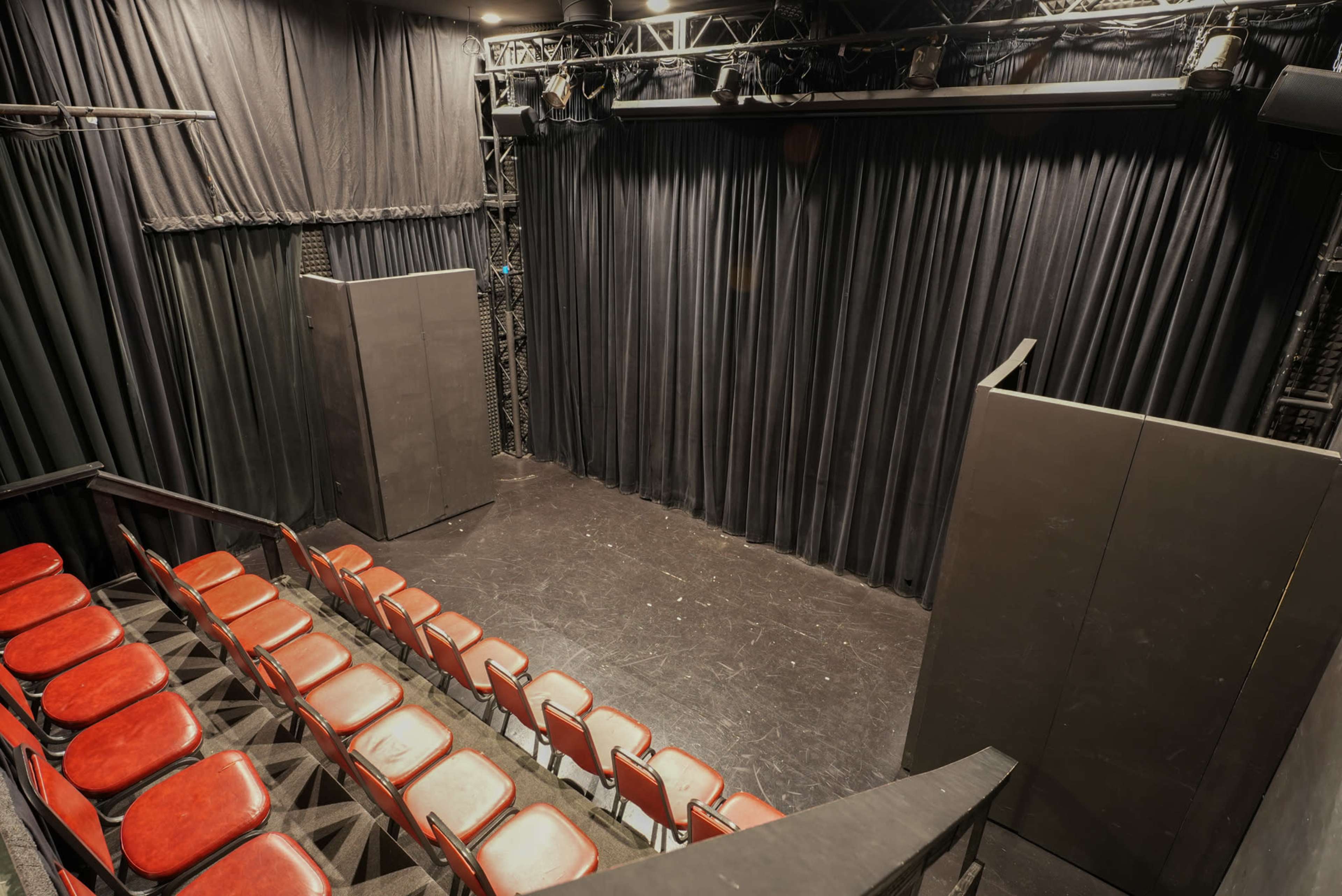 The image shows a small theater stage with black curtains and empty red seats arranged in a semi-circle.