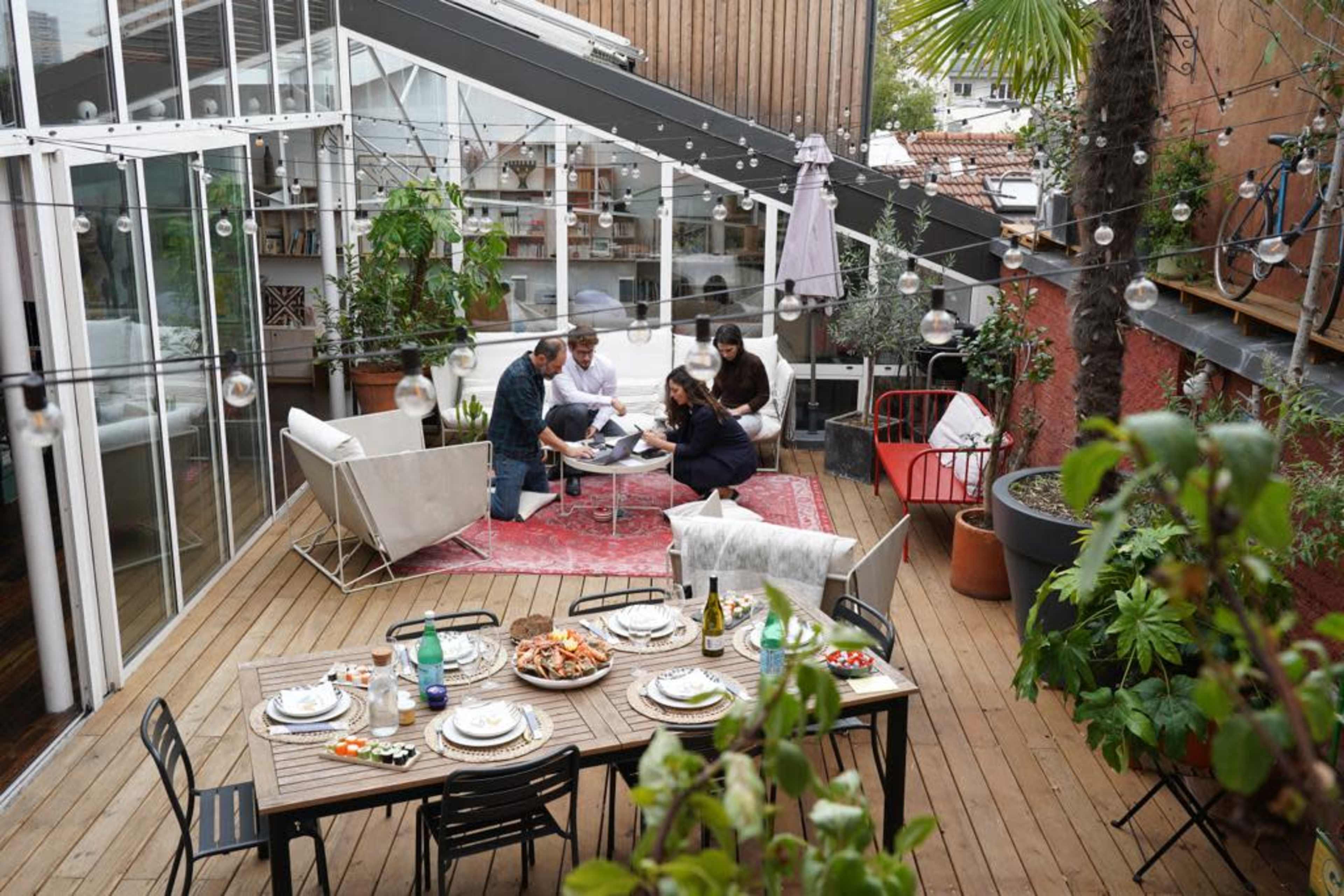 A group of five people gathers around a low table in an outdoor living space, surrounded by plants and string lights, with a dining table set for a meal nearby.