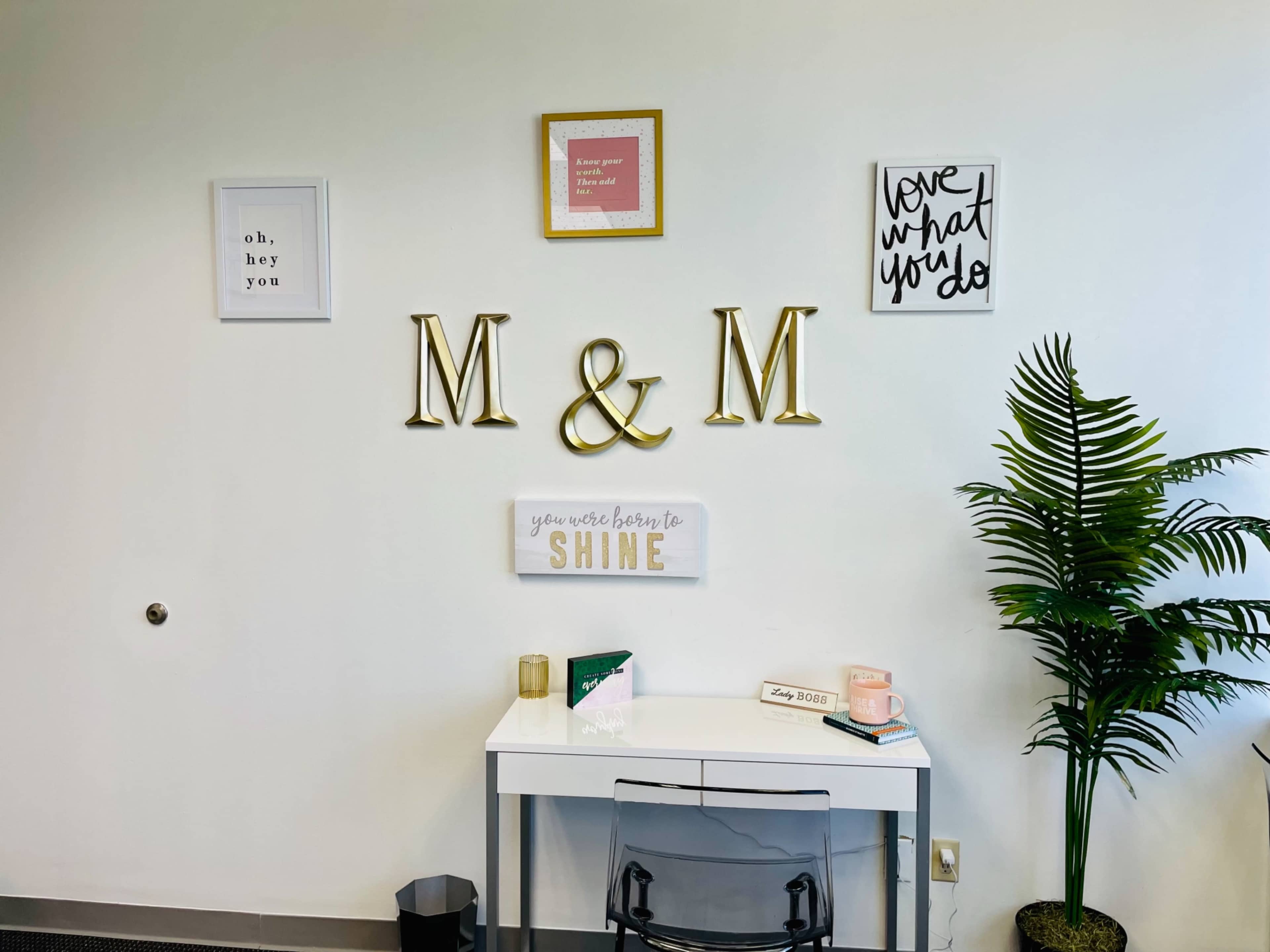 A minimalist workspace featuring a white desk, a clear chair, decorative wall art, and a small green plant in the corner.