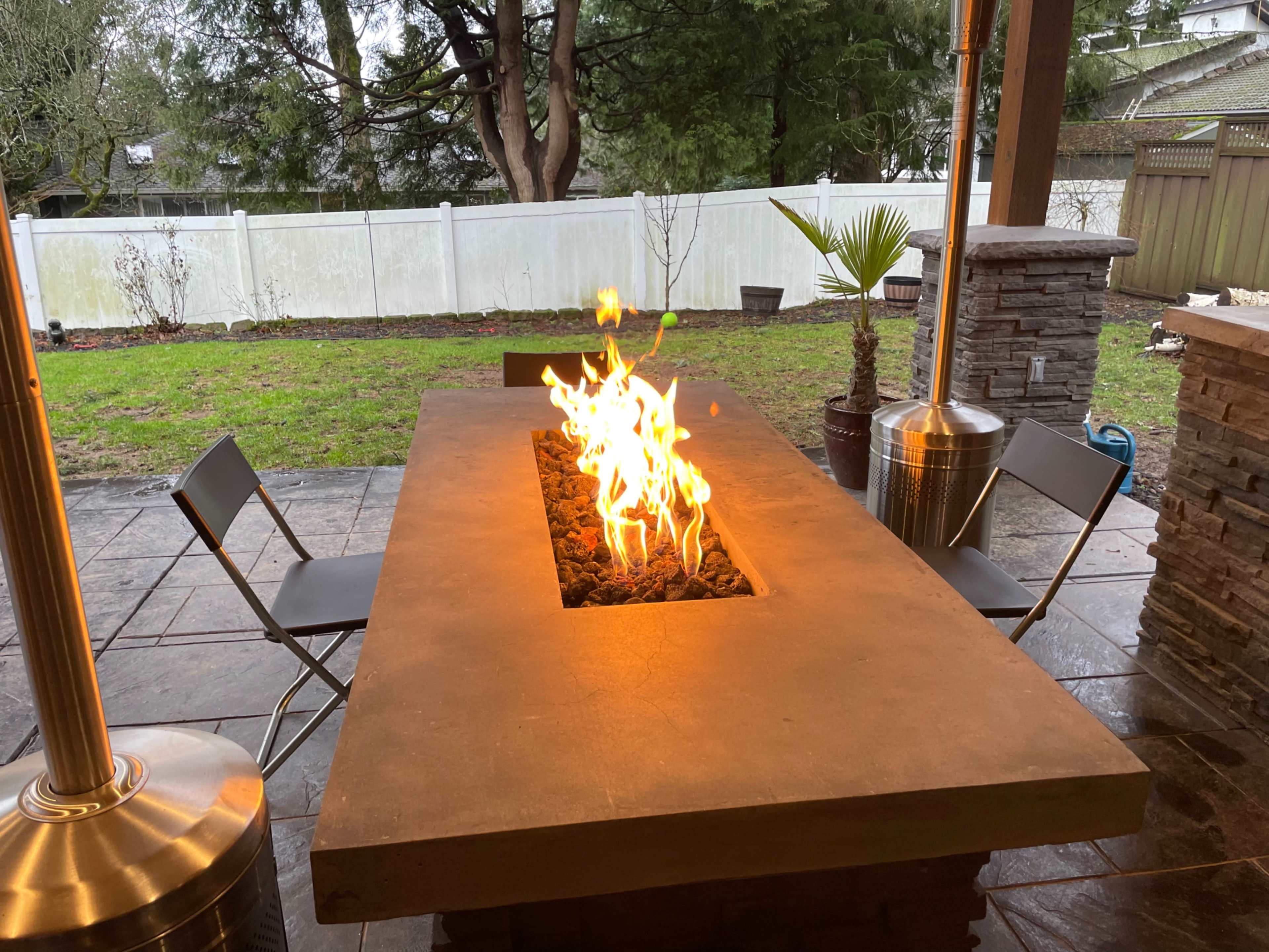 A modern stone fire pit table with flames rising from the center, surrounded by two metal chairs and a green yard in the background.