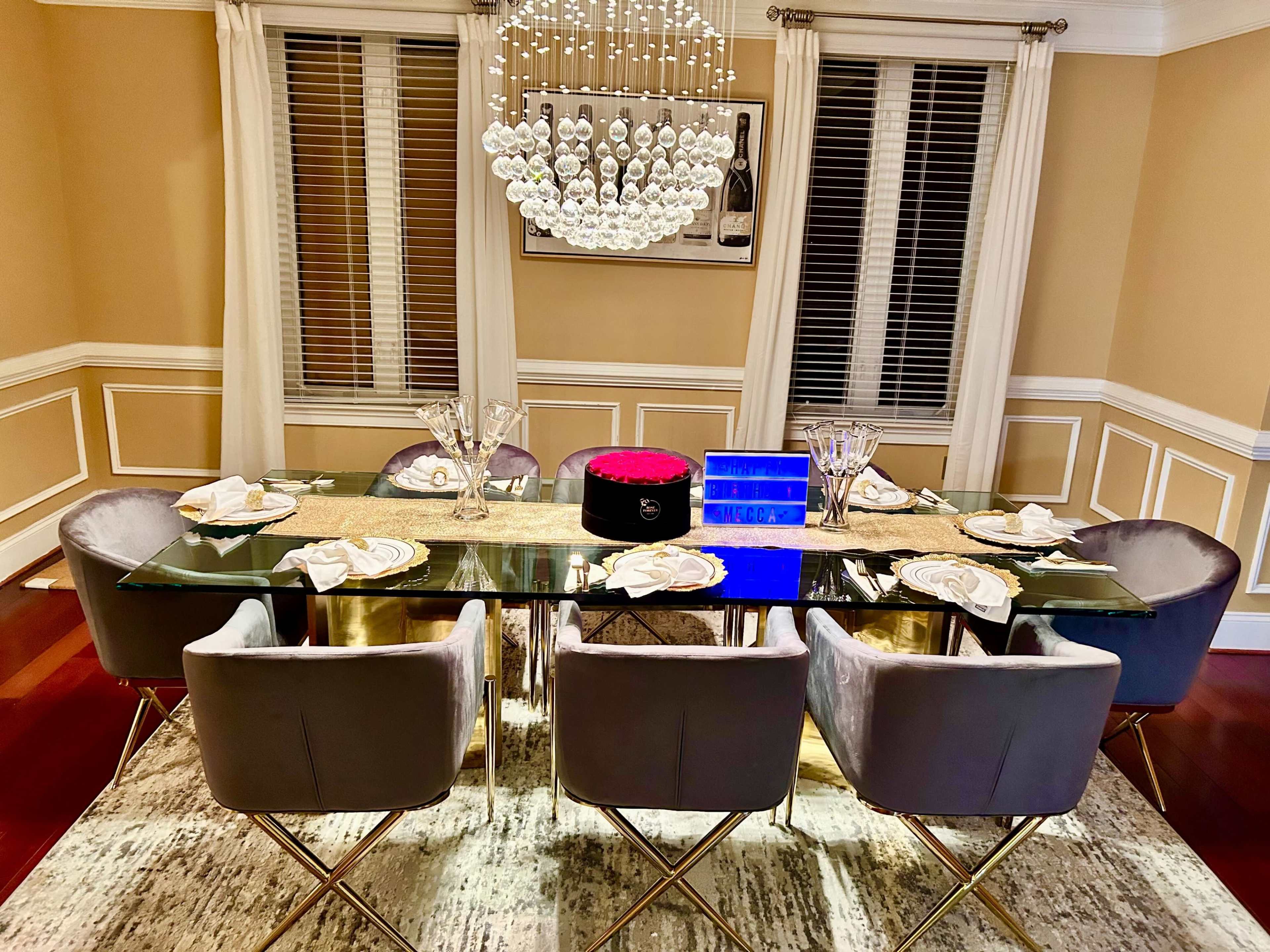 A glass dining table is set with elegant tableware and surrounded by upholstered chairs, beneath a modern chandelier in a well-decorated dining room.