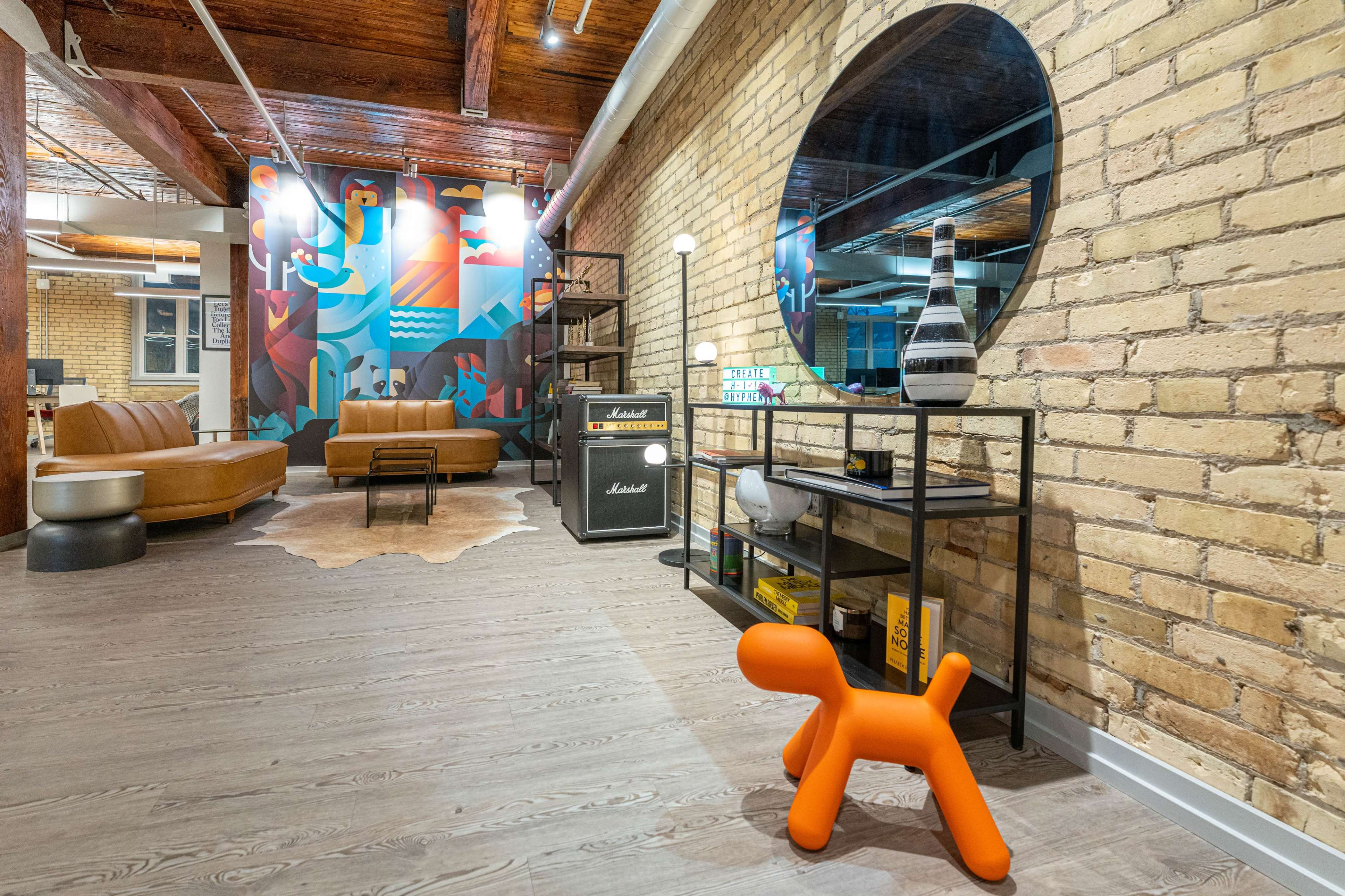 A modern living space with a brick wall, a large round mirror, a stylish black shelf with decor items, two brown leather sofas, and a playful orange dog-shaped sculpture.