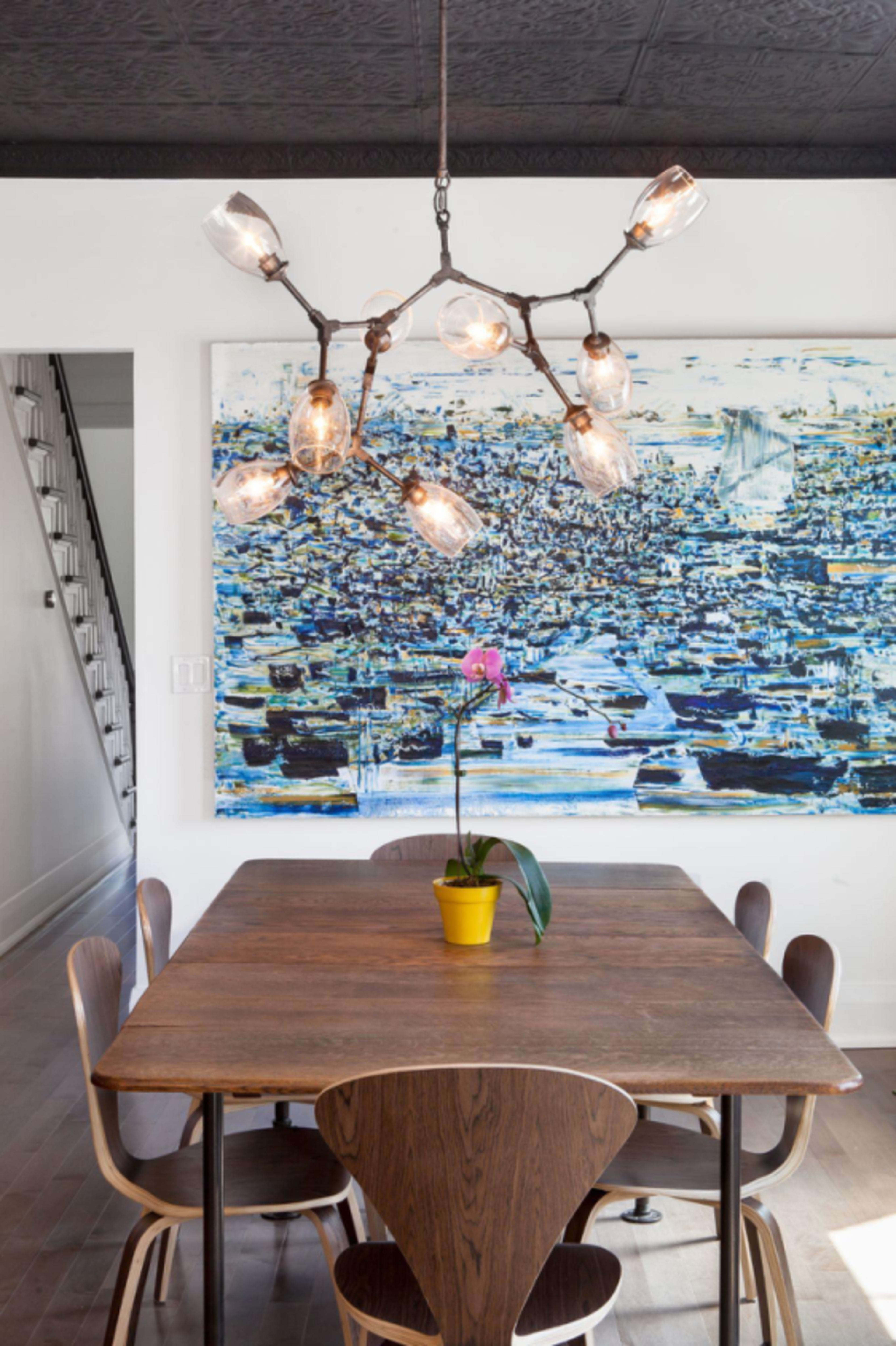 A wooden dining table with a potted orchid centers a room featuring a unique pendant light and a large abstract painting on the wall.