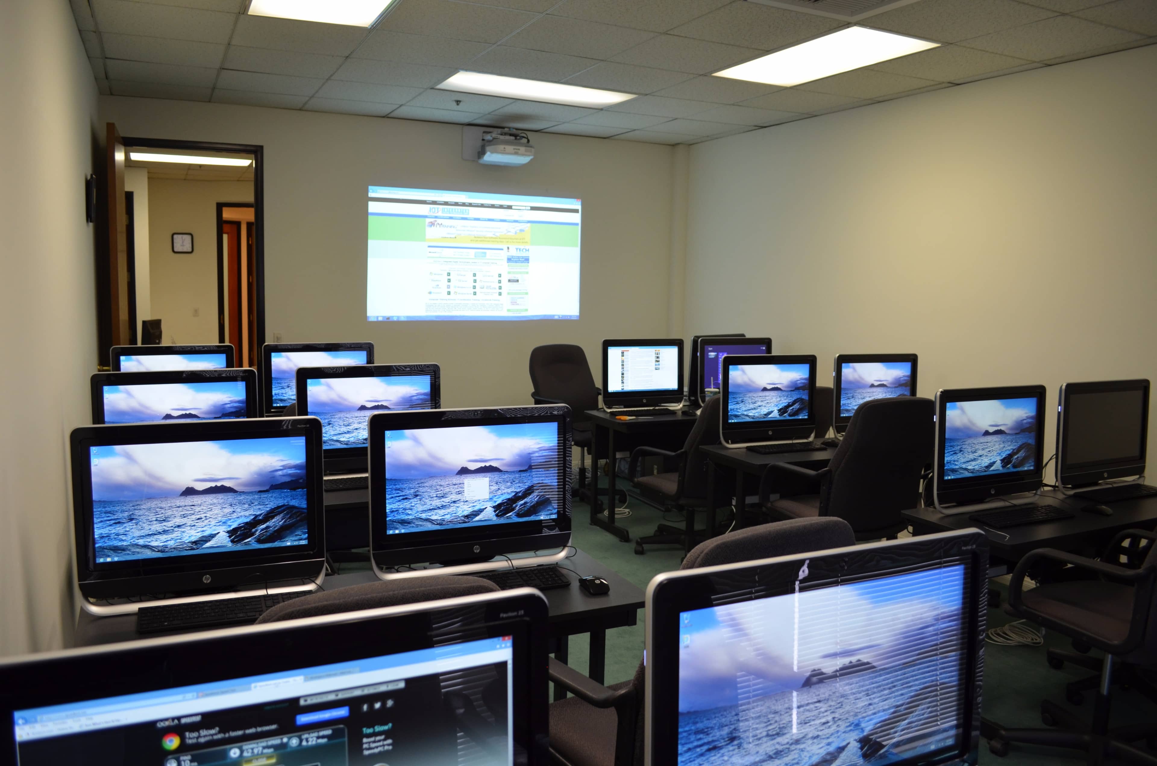 A computer lab is equipped with multiple desktop computers arranged in rows, each displaying images of ocean landscapes, and a projector displaying a webpage on the wall.