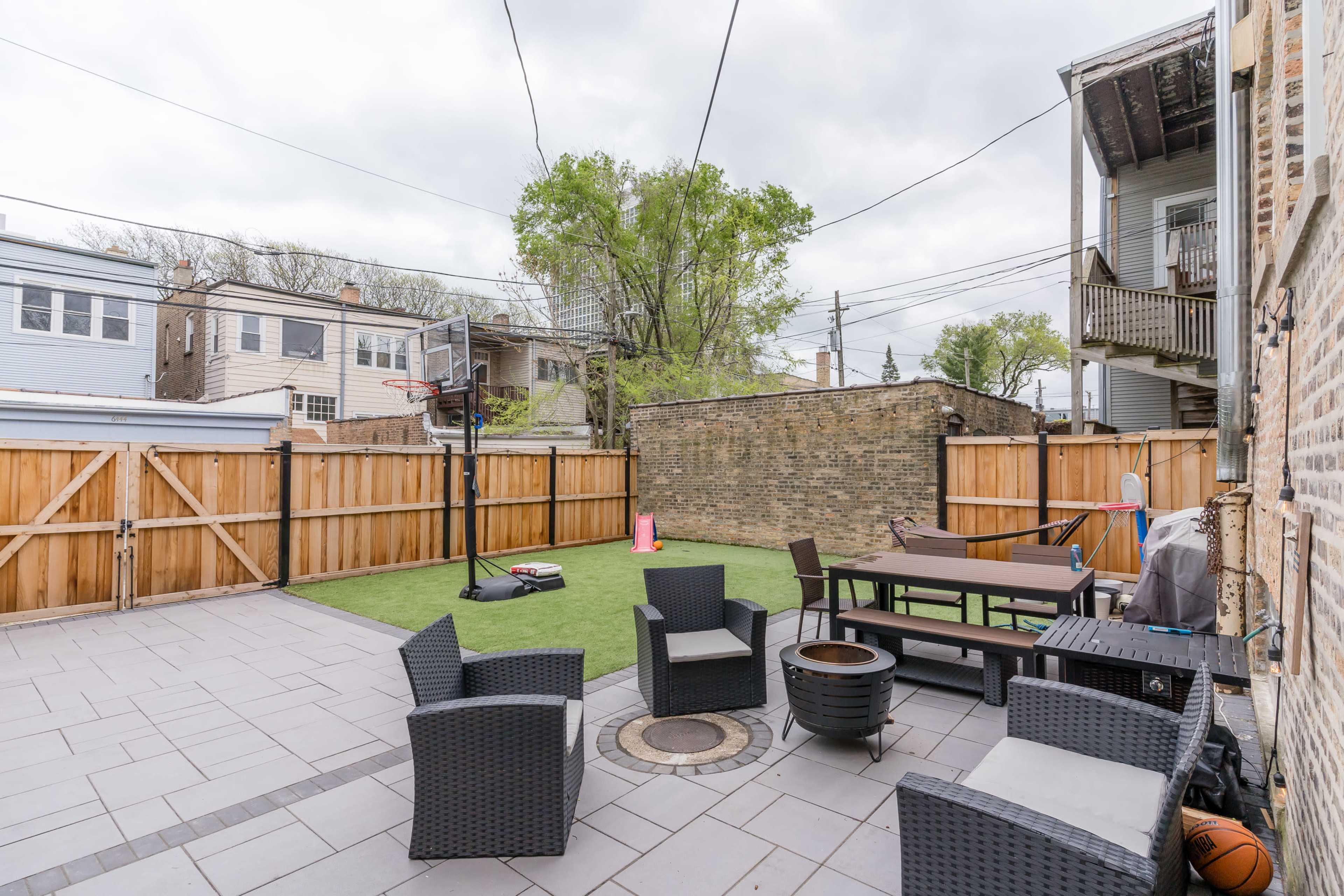 The image shows a spacious backyard with a basketball hoop, a seating area made of dark wicker furniture, and a paved section surrounded by wooden fencing.