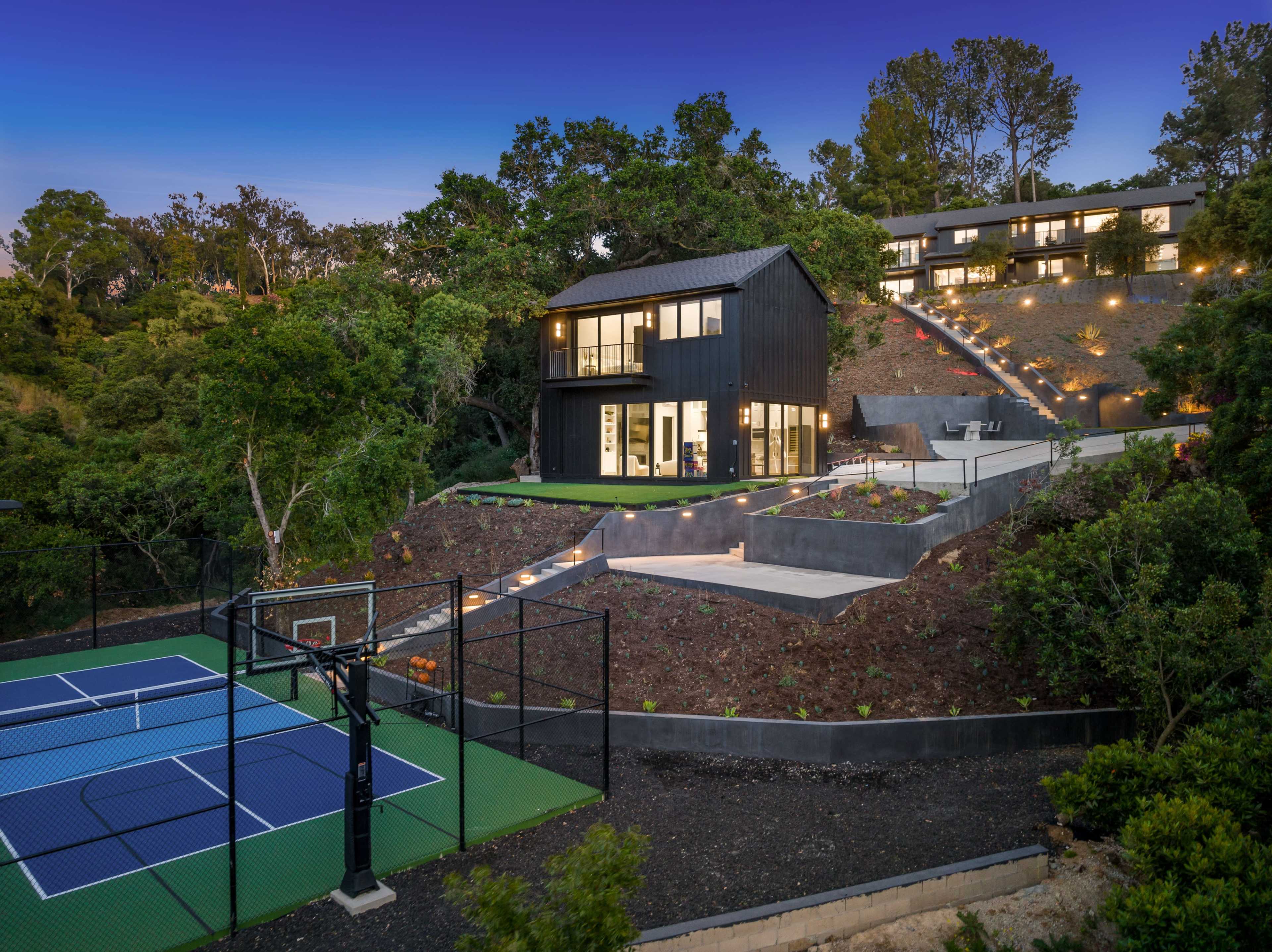 A modern black house with large windows is situated on a sloped lot, featuring a basketball court in the foreground and terraces planted with greenery in the background.