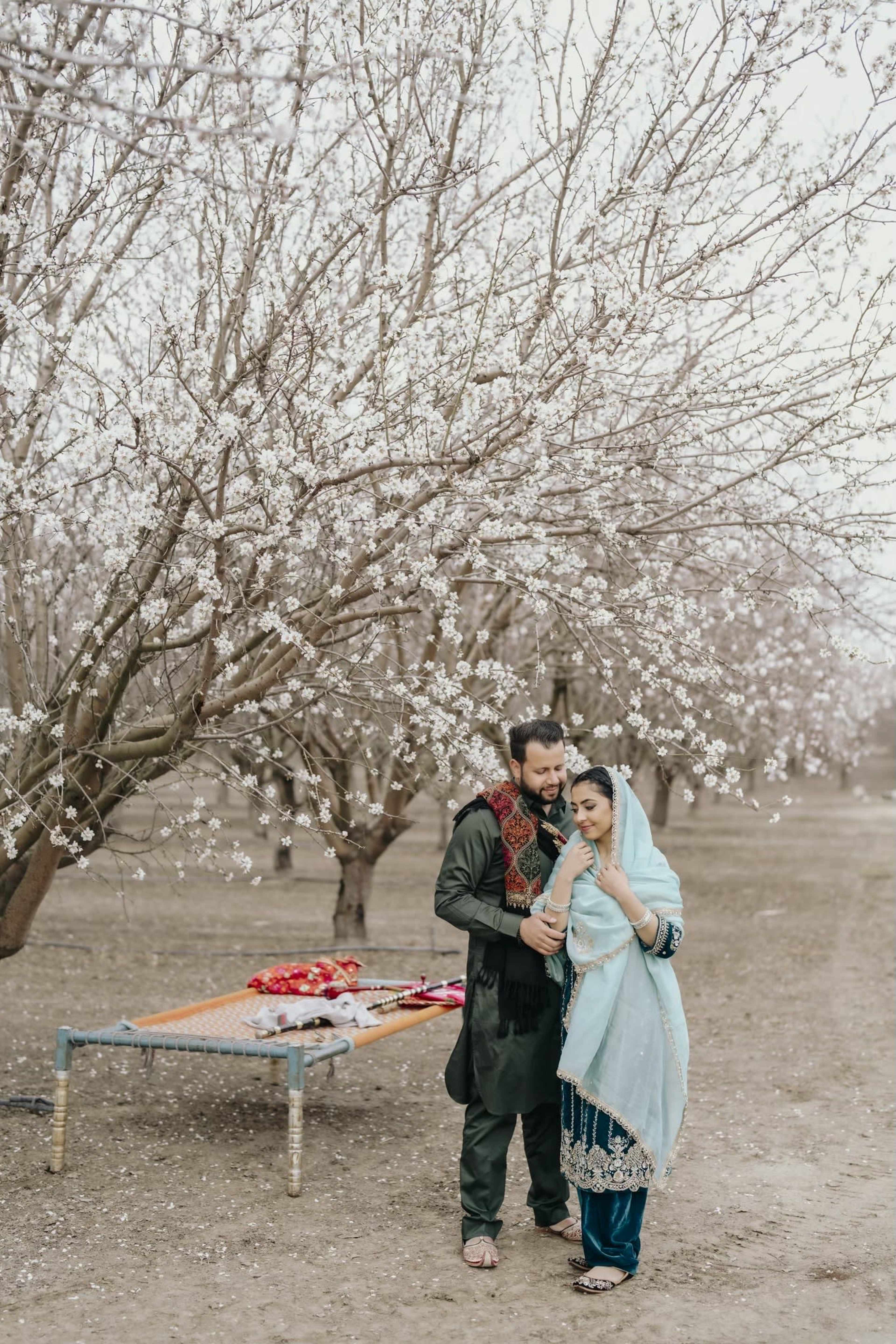 A couple stands close together among blooming trees, with a bed of flowers and a traditional picnic setup in the background.