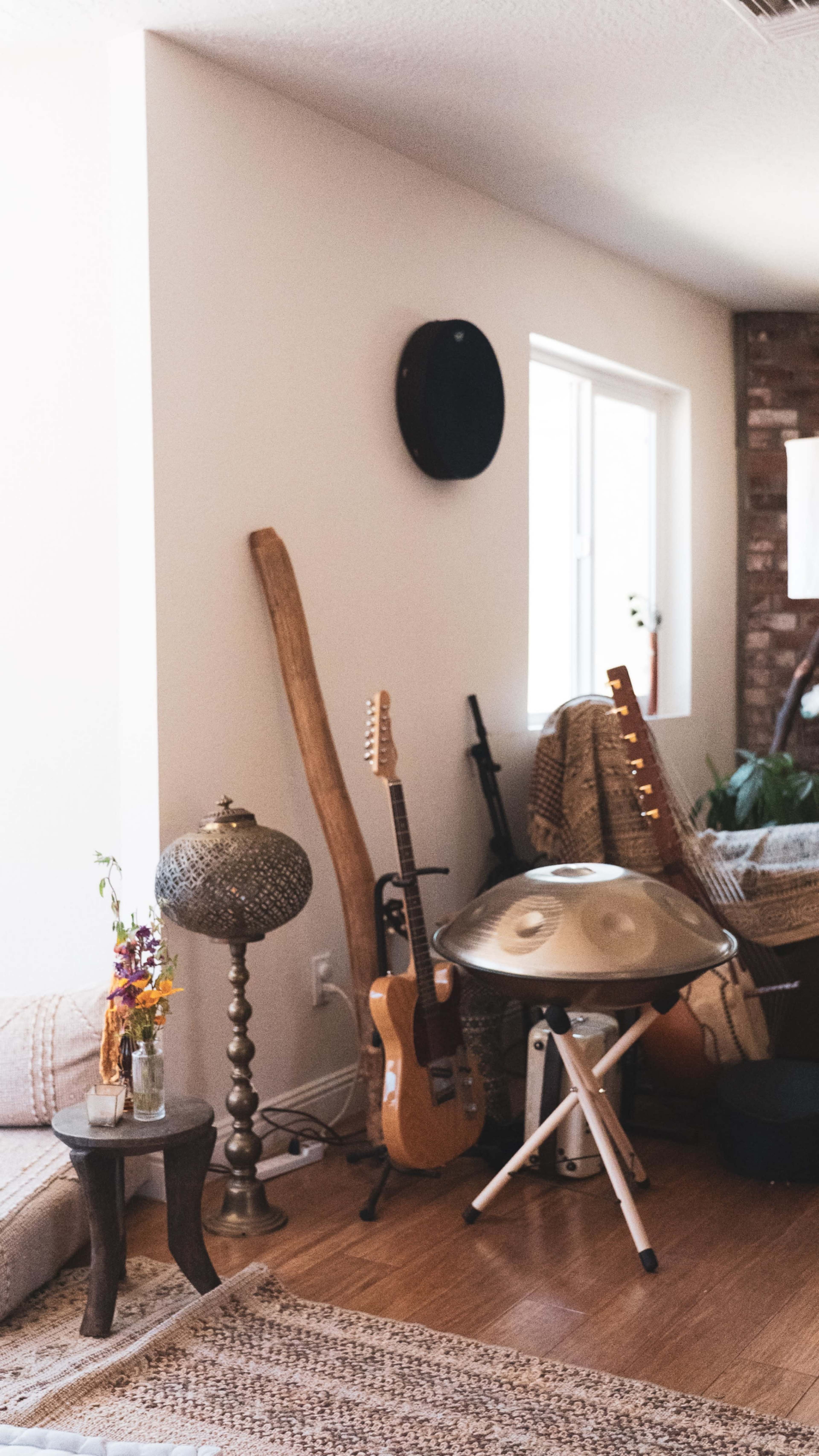 The image shows a cozy living space featuring a collection of musical instruments, a decorative lamp, and a small table alongside a couch and a window.
