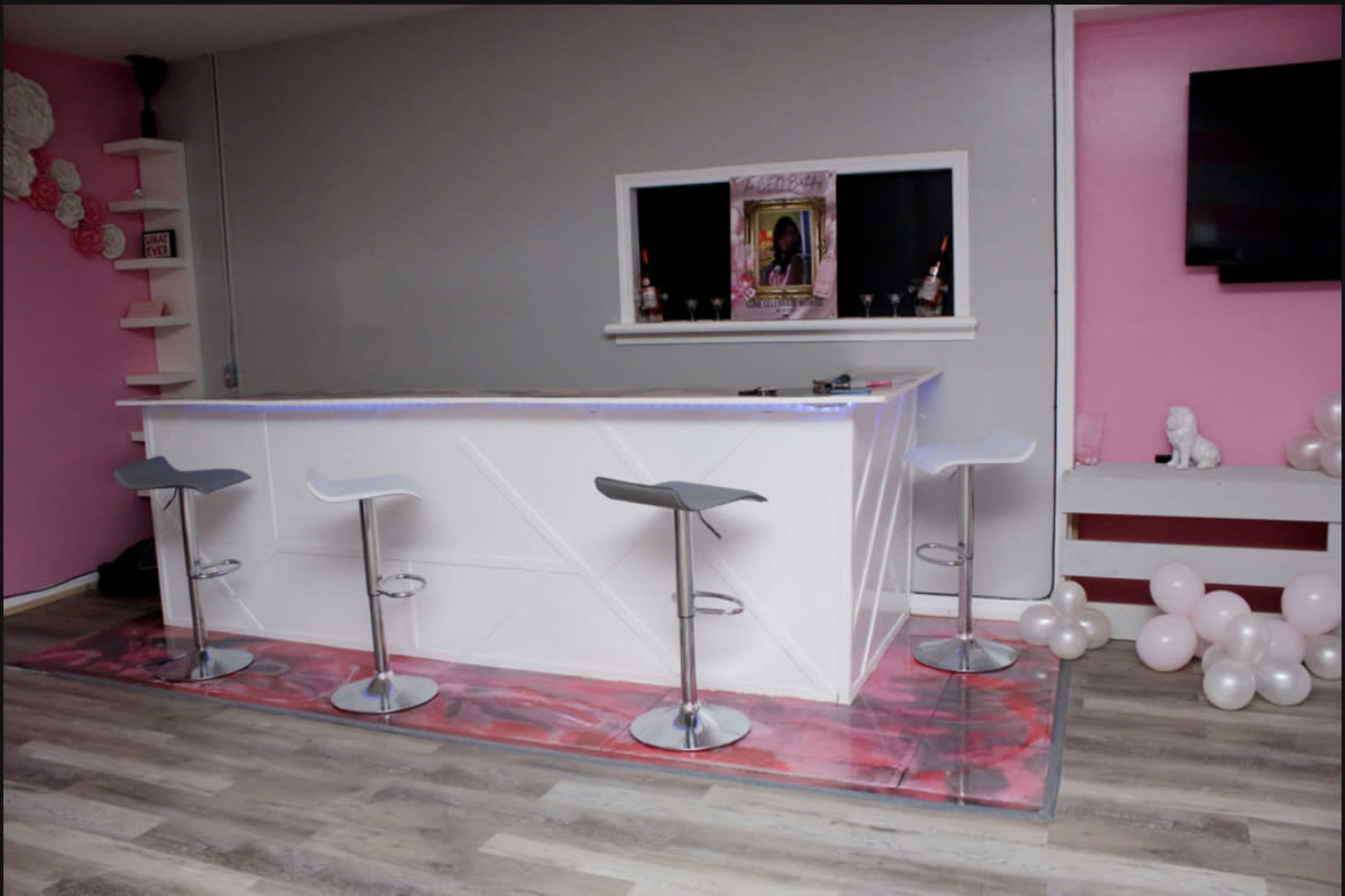A modern bar area features a white countertop with four chrome stools, accented by pink walls and decorative elements.