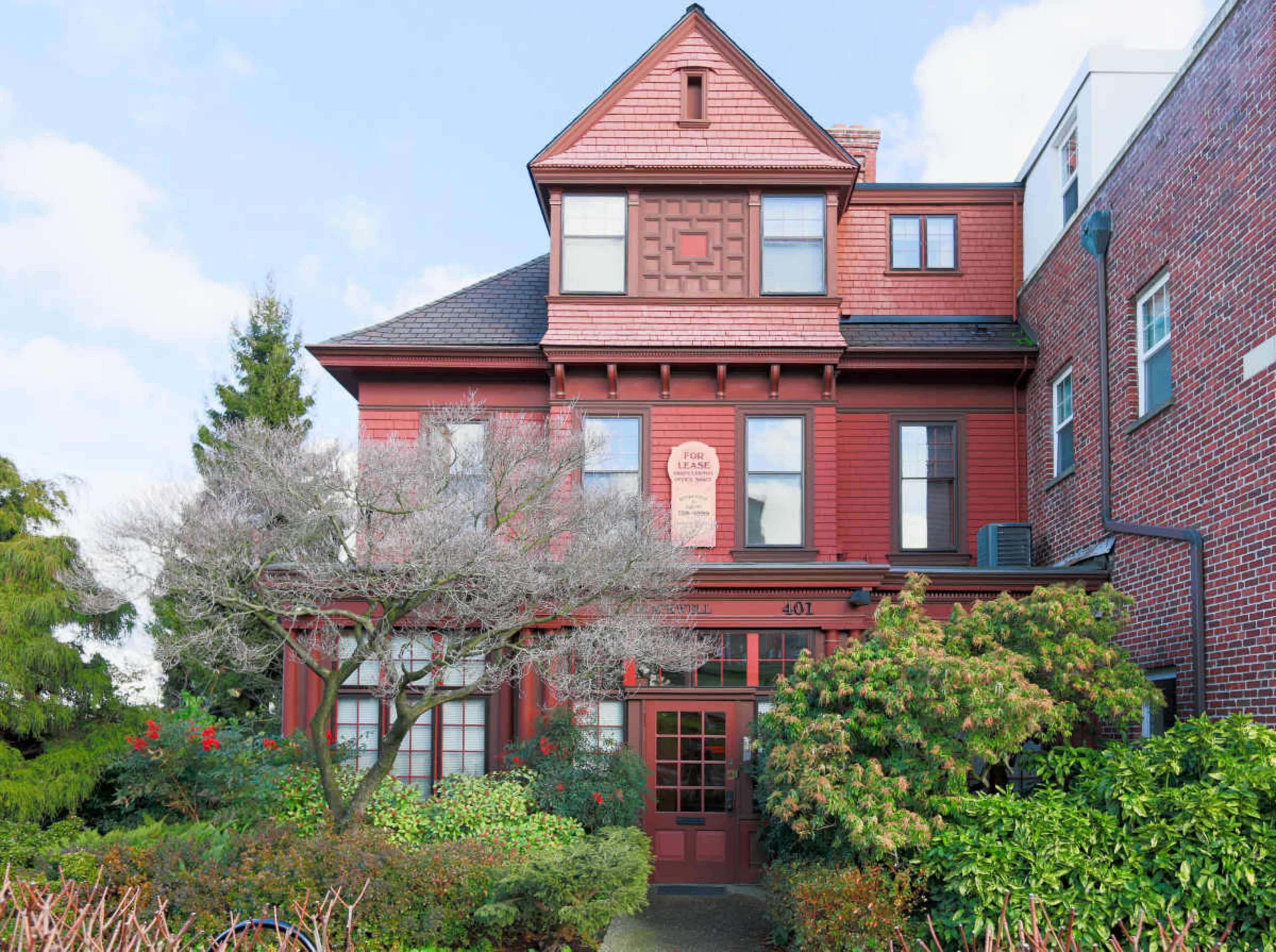 The image shows a large, red Victorian-style house with a peaked roof and elaborate architectural details, surrounded by green shrubs and a flowering tree.