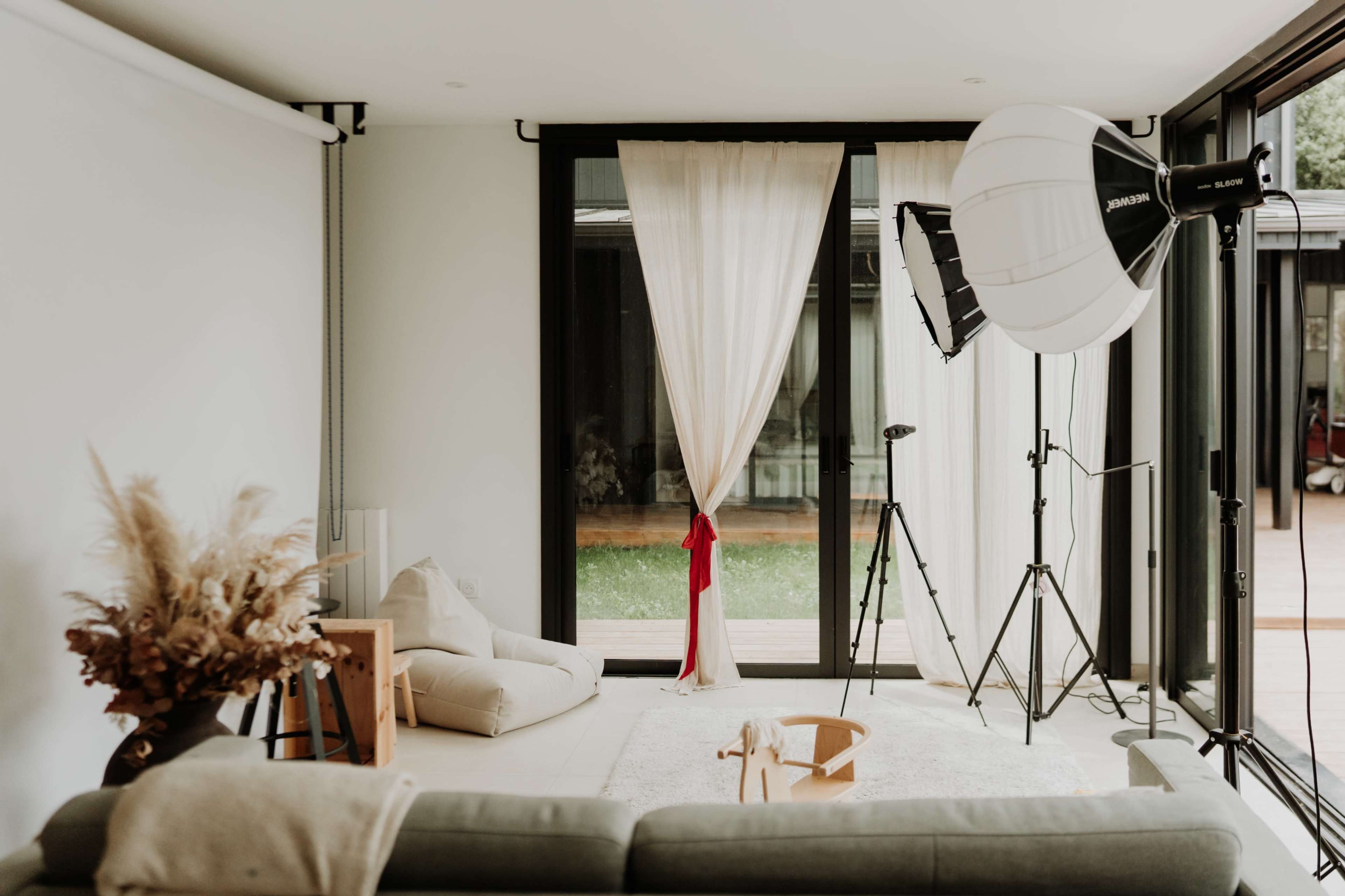 The image shows a bright and spacious room with large windows, a soft couch, a photography backdrop, and studio lighting equipment.