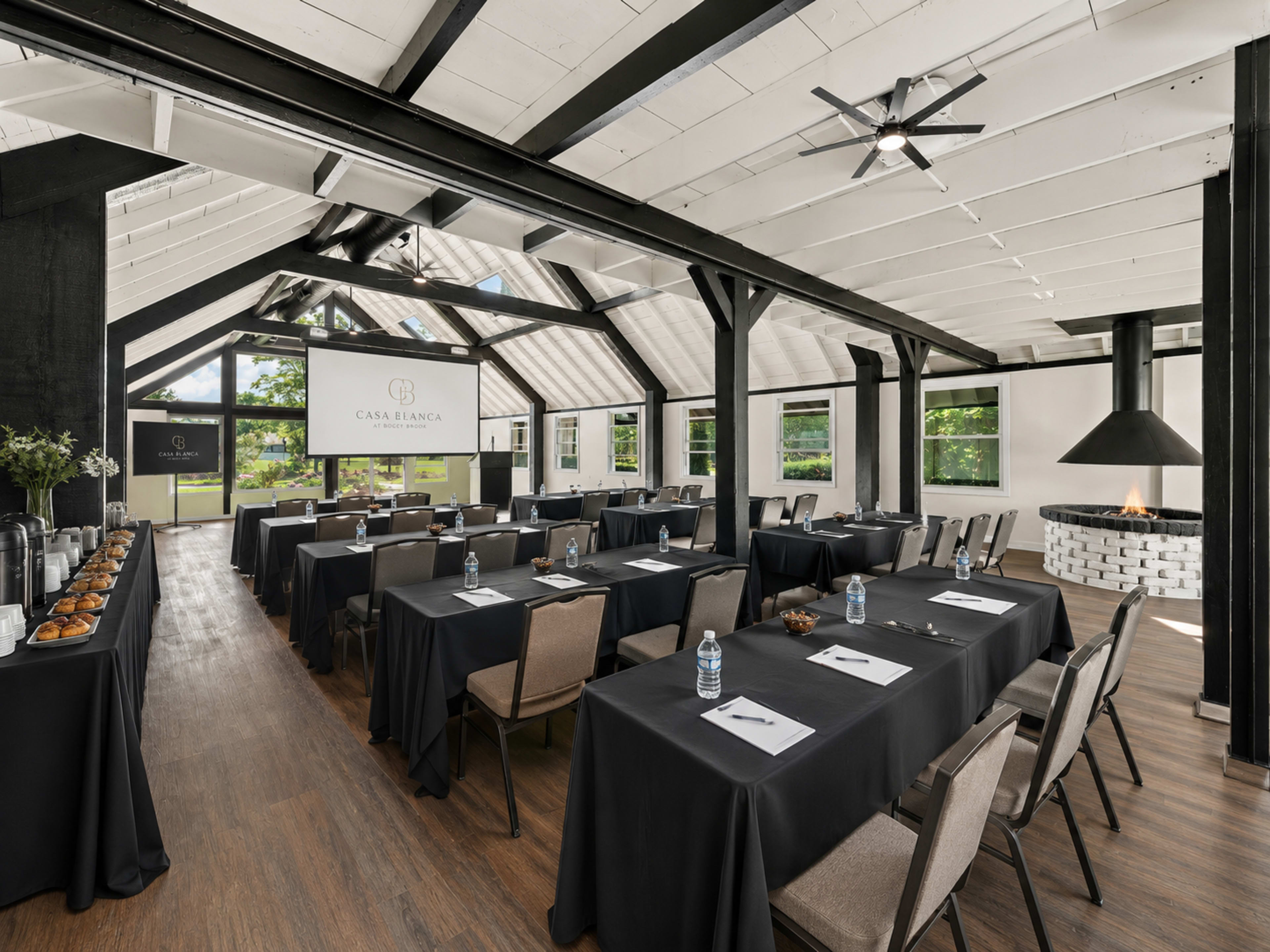 The image shows a well-lit meeting room with rows of tables covered in black tablecloths, featuring a projector screen at the front and a modern fireplace in the corner.