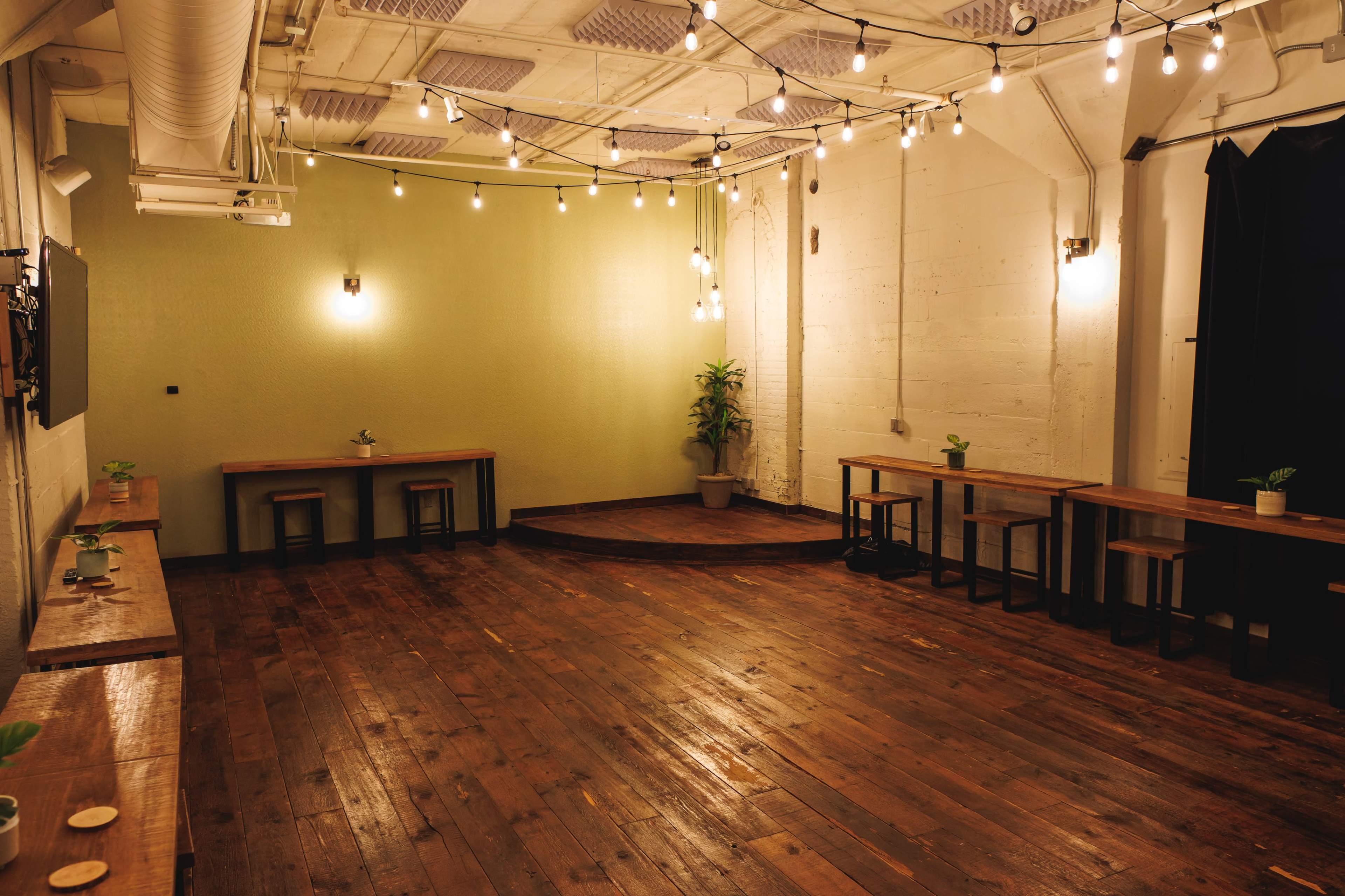 A spacious room with wooden floors, equipped with several wooden tables, plants, and overhead string lights.