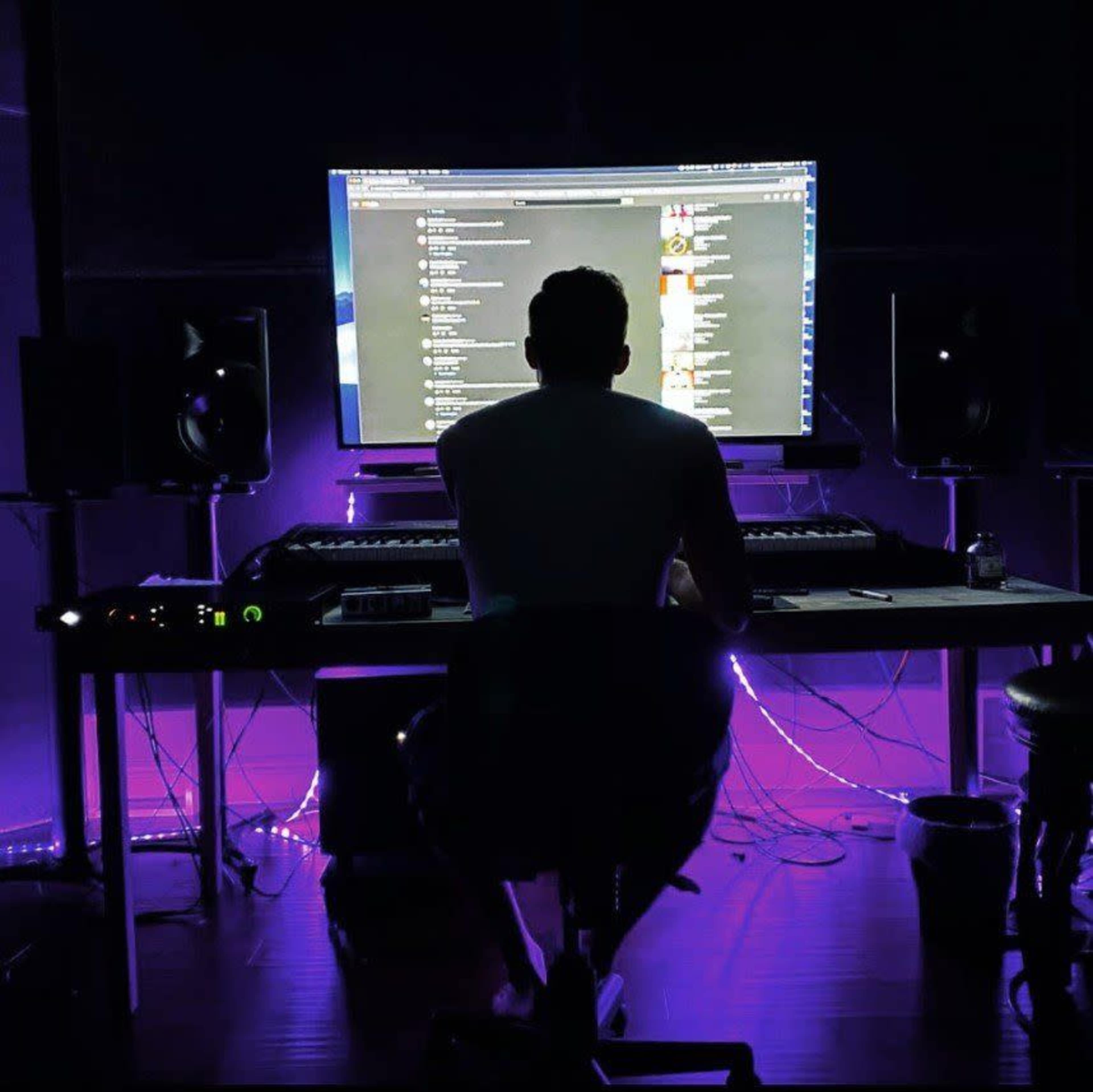 A person sits at a desk in a dimly lit room, facing a large monitor with music software visible.