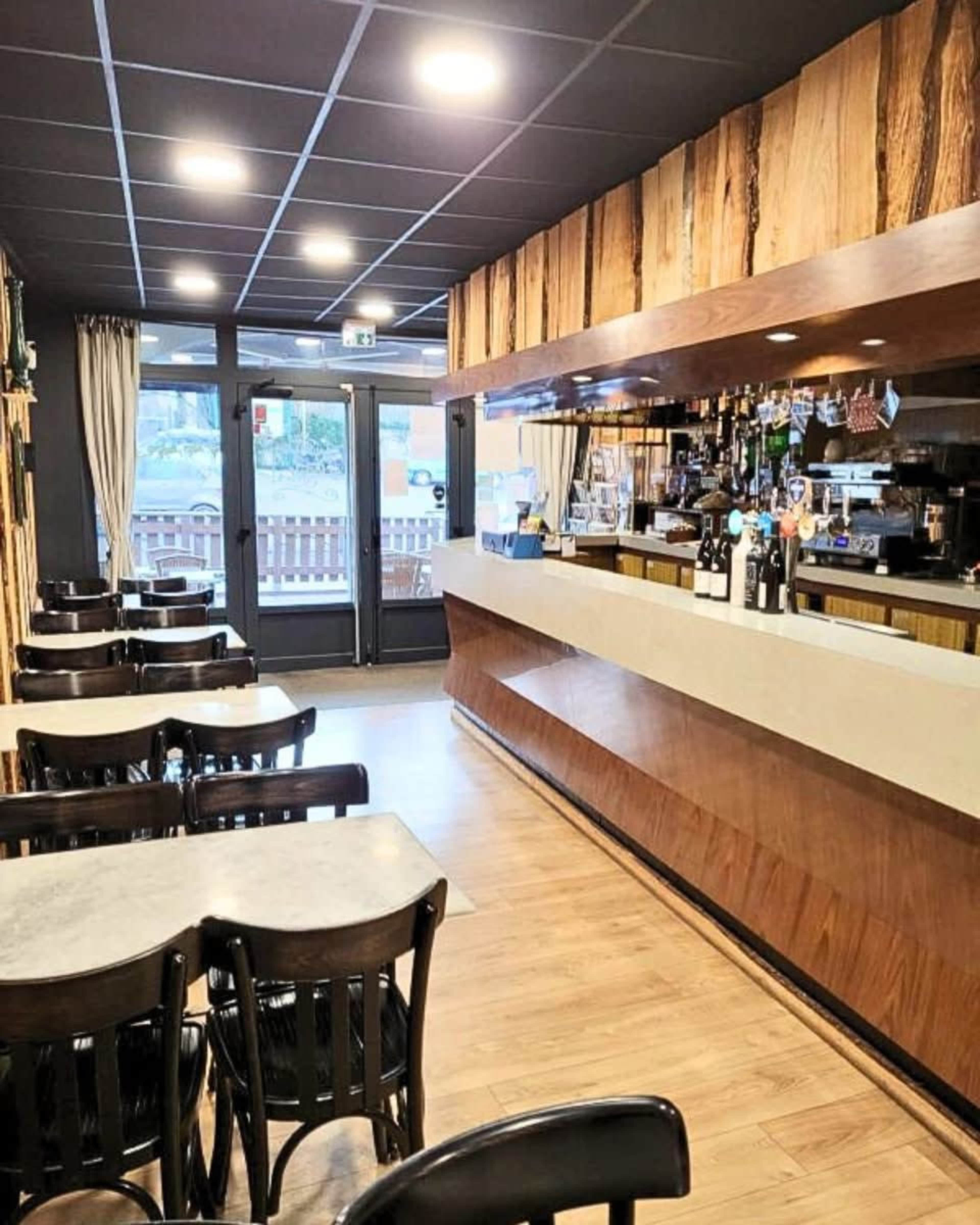 The image shows the interior of a restaurant or bar with wooden decor, a counter featuring drinks, and empty tables arranged neatly.