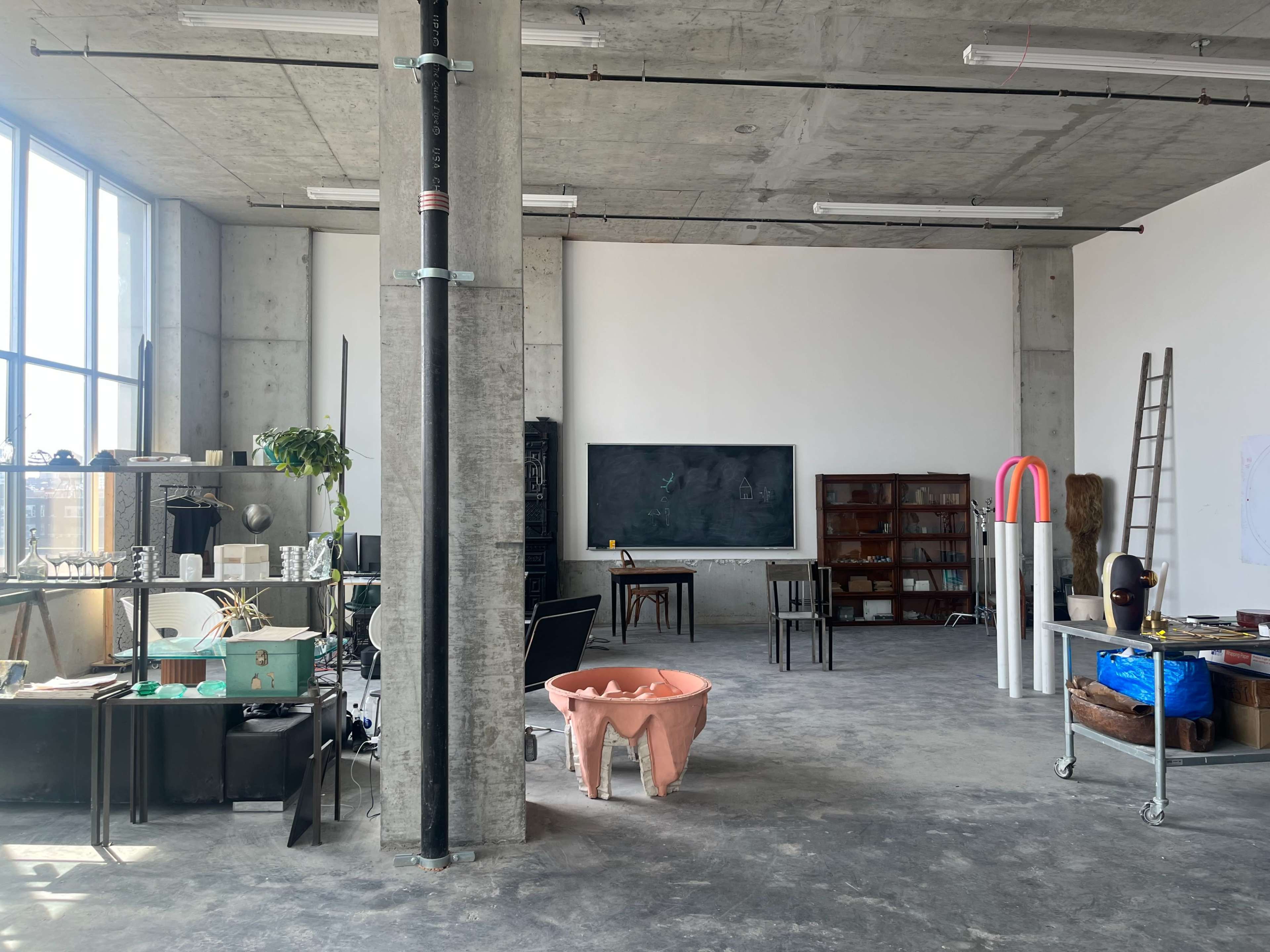 The image shows a spacious, industrial-style room with concrete walls, various furniture items, and large windows allowing natural light to enter.