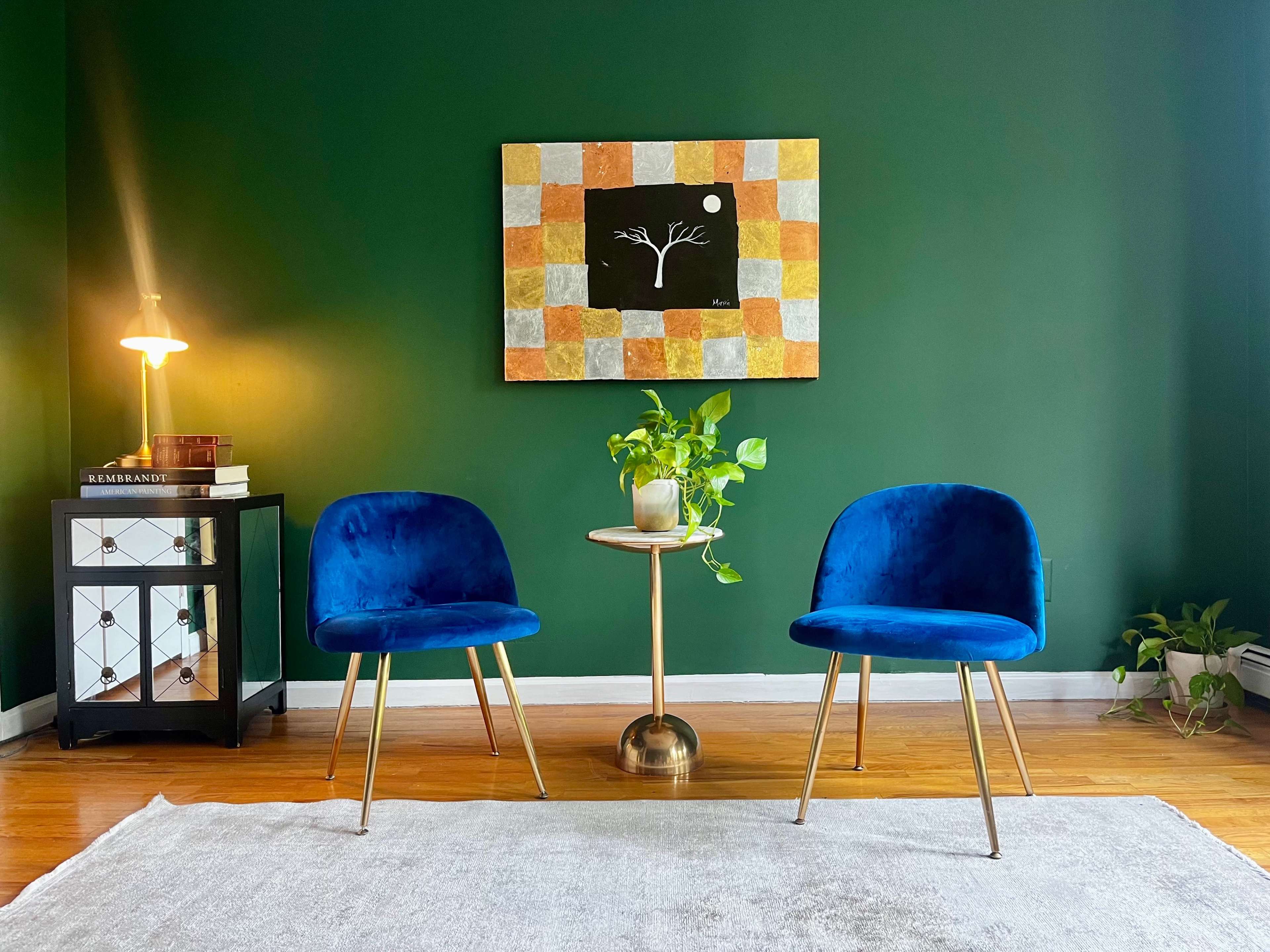 A cozy room with two blue chairs, a small round table with a plant, a lamp, and a painting on a dark green wall.