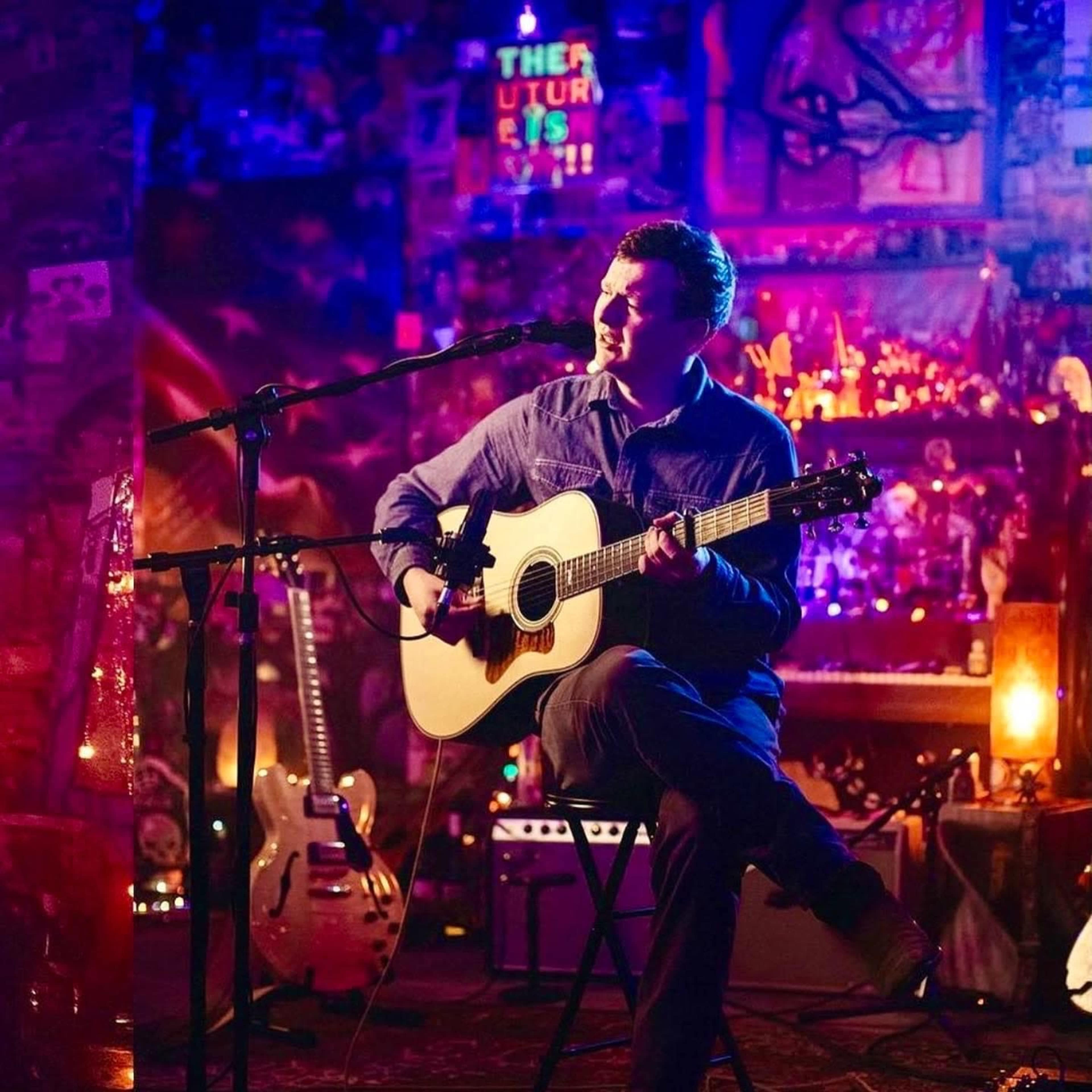 A musician sits on a stool performing with an acoustic guitar in a dimly lit venue filled with colorful decorations.