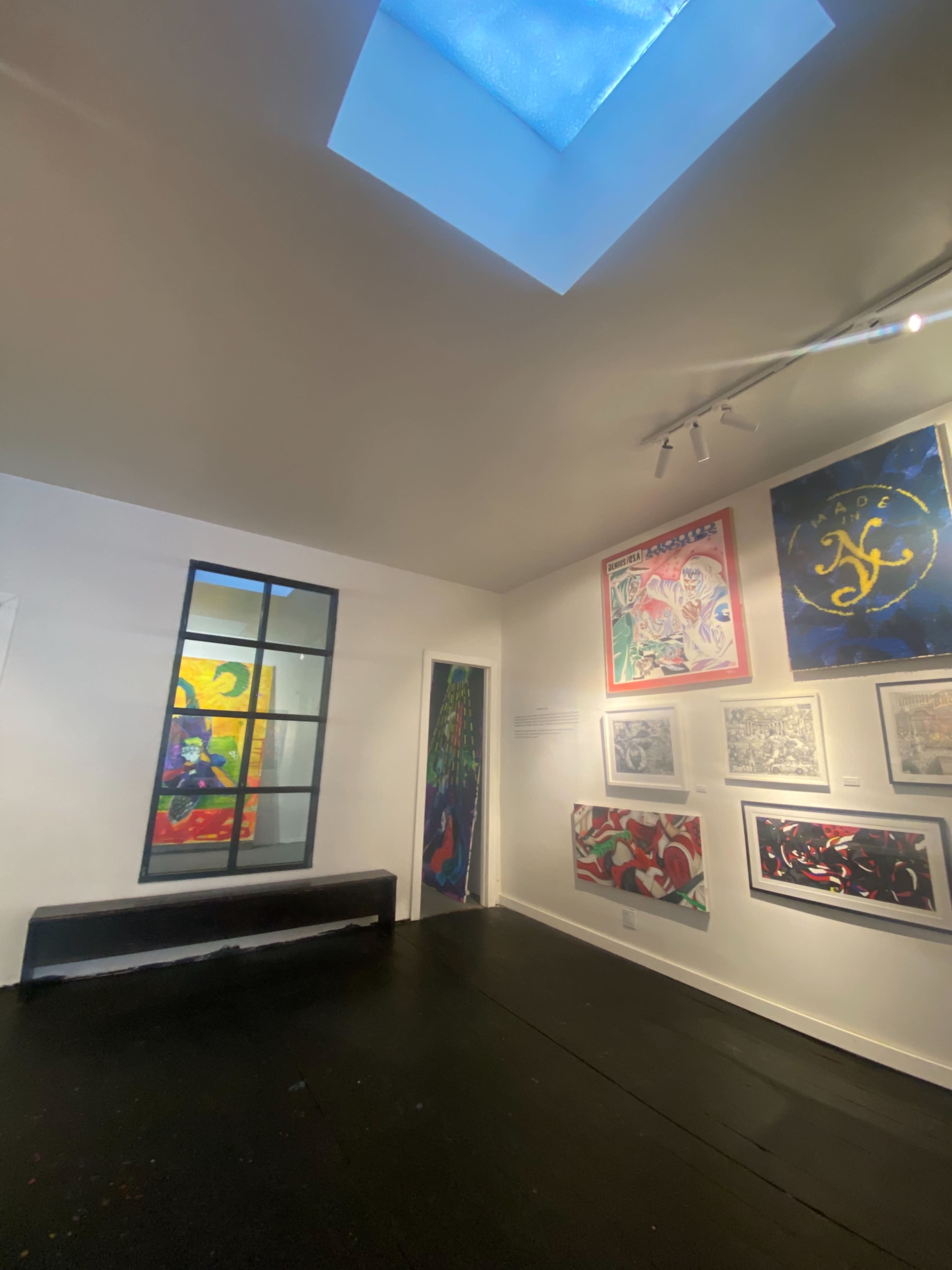 The image shows an art gallery with colorful wall artworks, a skylight, and a black bench against the wall.
