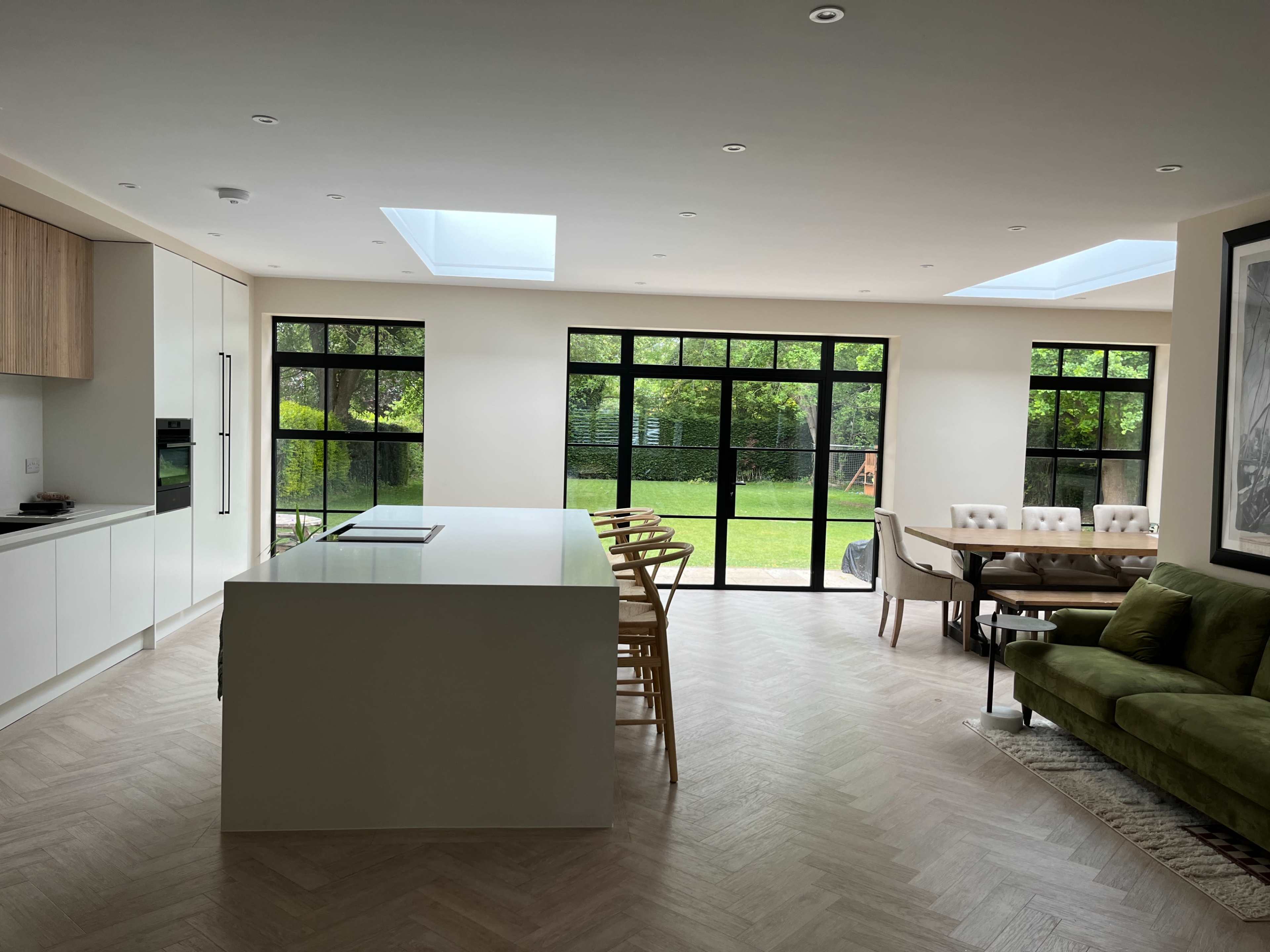 The image shows a modern open-plan kitchen and dining area with large windows overlooking a green garden.