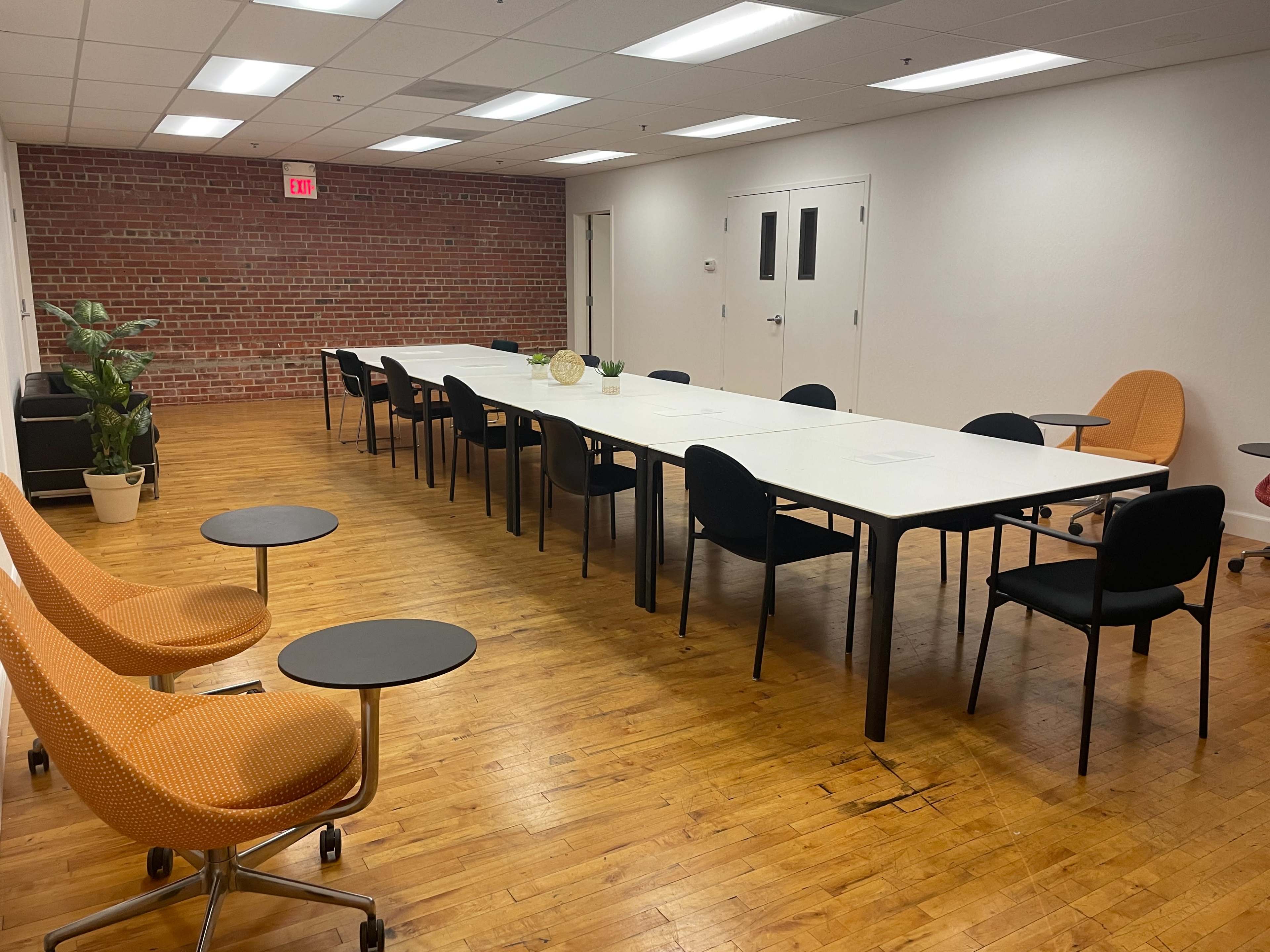 A spacious conference room features a long white table surrounded by black chairs, a few small side tables, and a potted plant, with a brick wall in the background.