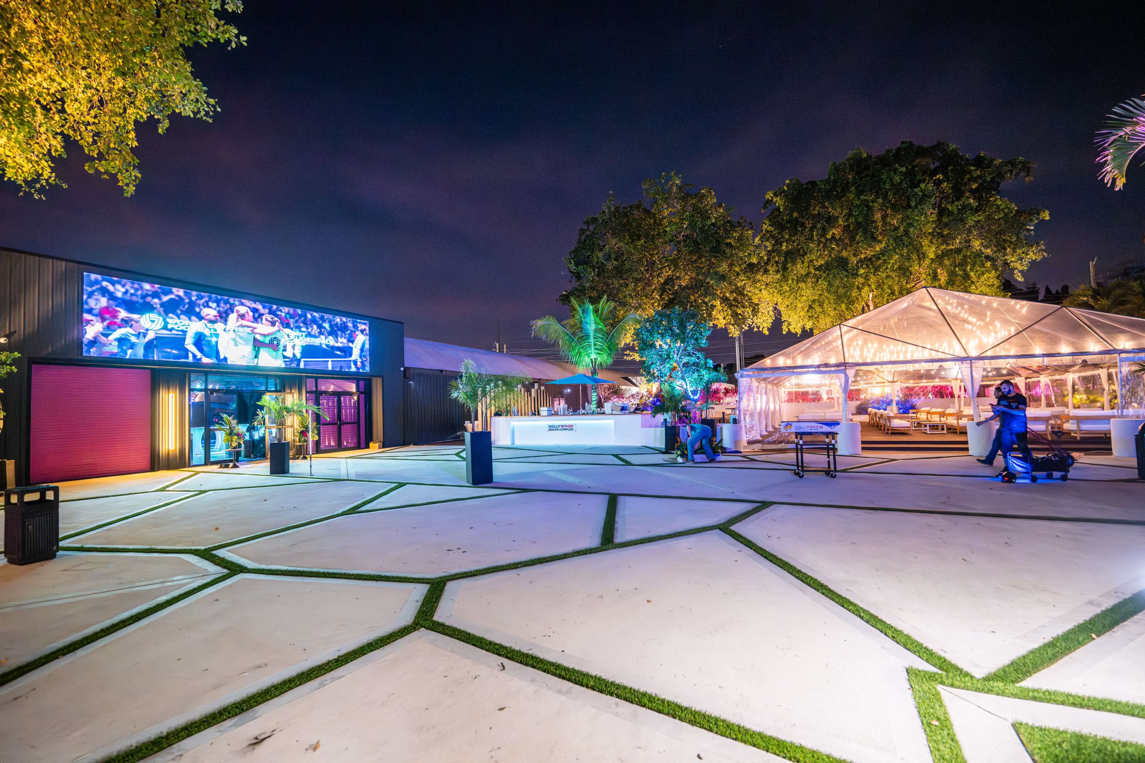 The image shows an outdoor event space at night, featuring a large clear tent illuminated with lights, a big screen displaying video, and geometric paving patterns on the ground.
