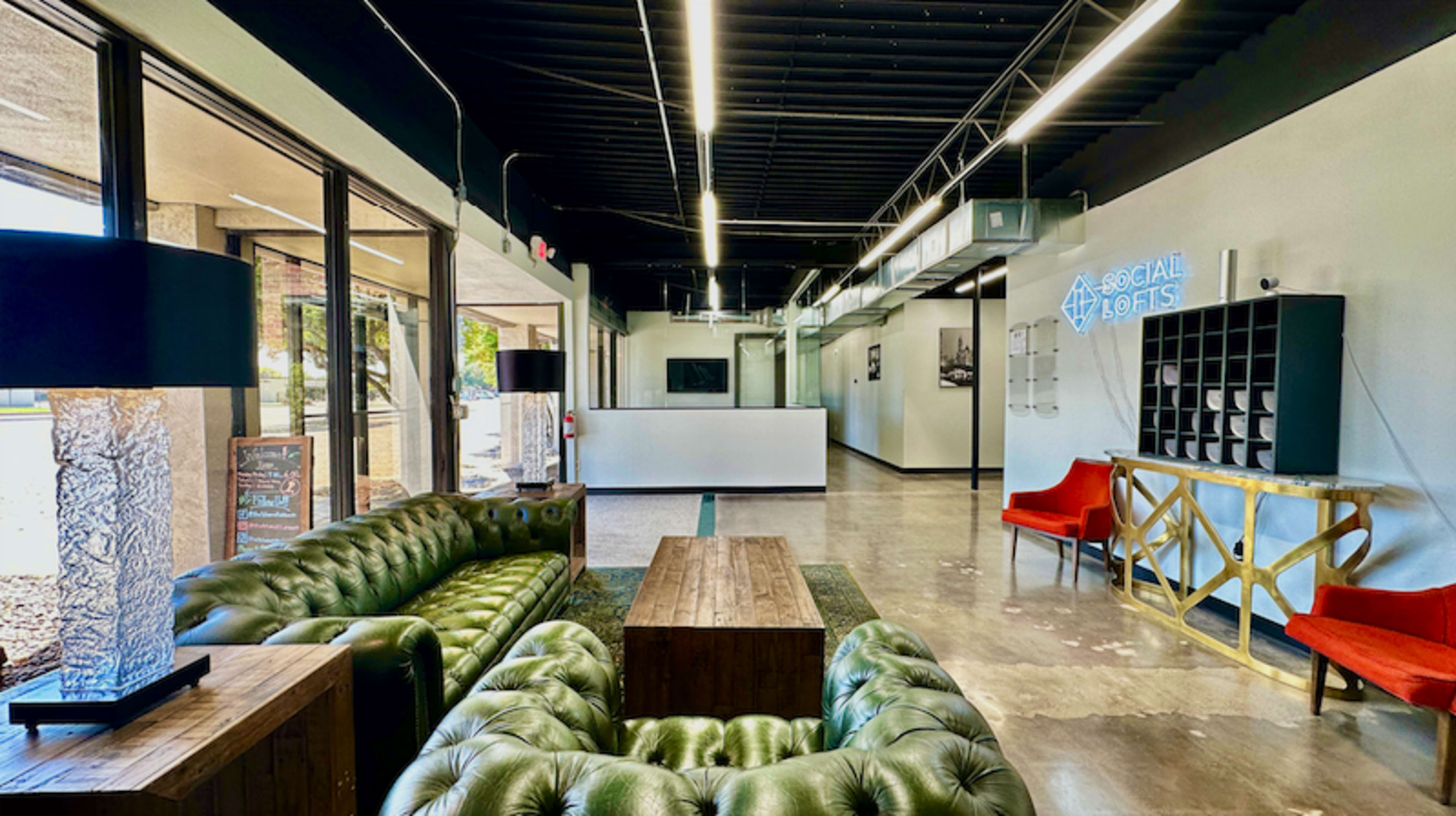 The image shows a modern lounge area featuring green leather sofas, a wooden coffee table, and red chairs, with large windows and a blue neon sign in the background.