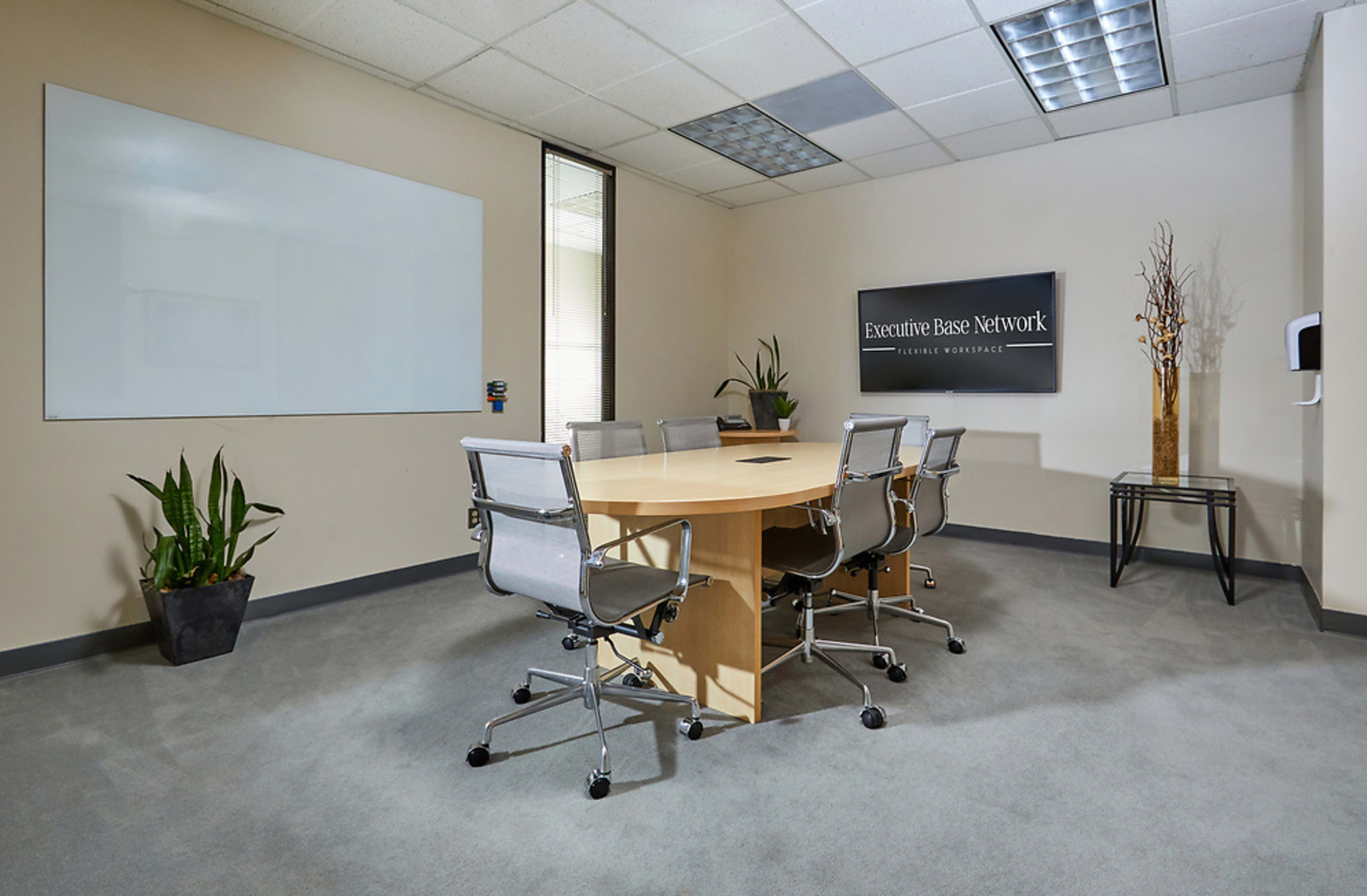A conference room featuring a round table surrounded by six metallic chairs, with a whiteboard on one wall and potted plants in the corners.